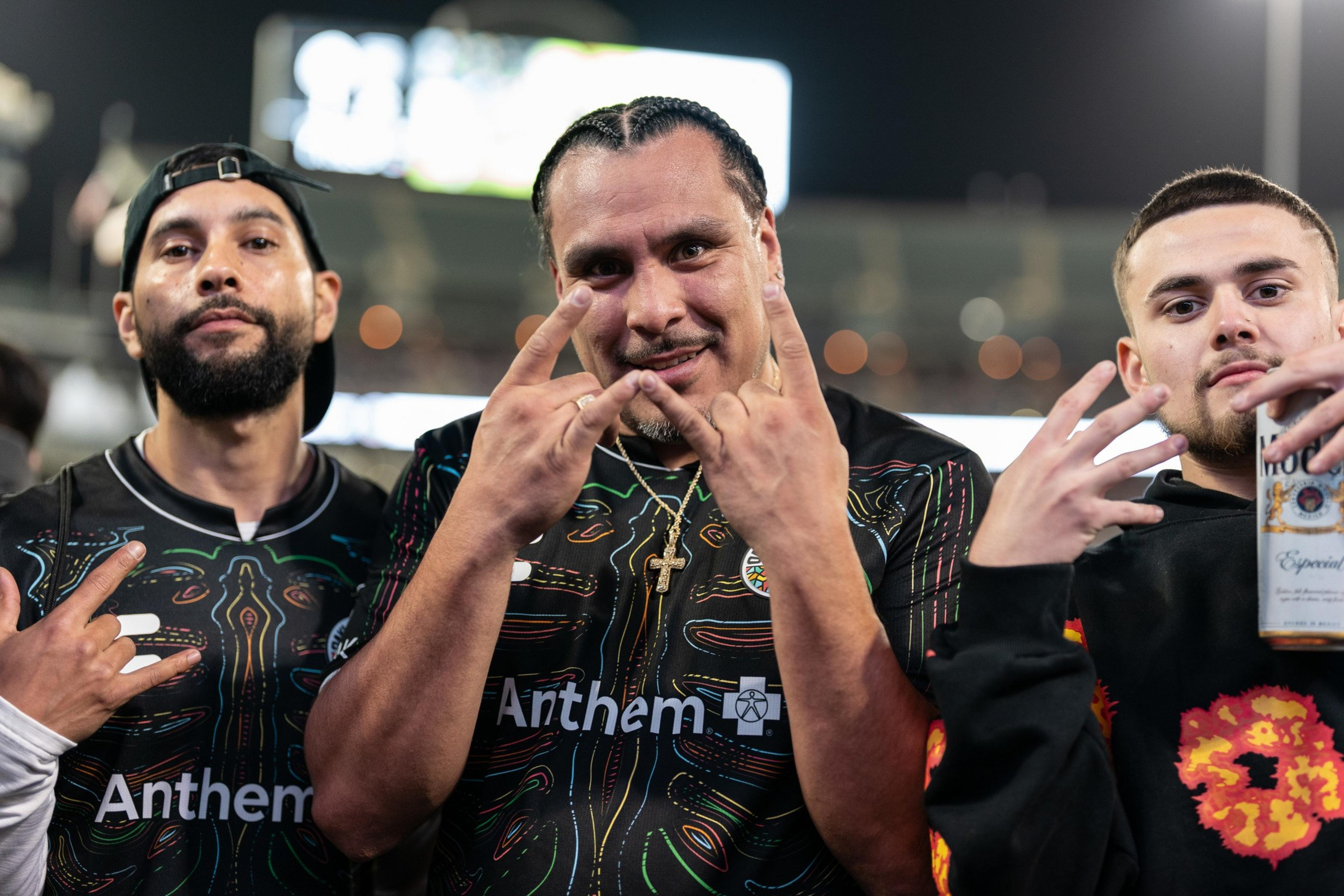 Three men stand close together at a nighttime sports event, wearing jerseys. They make hand gestures, and one holds a drink can, expressing excitement.