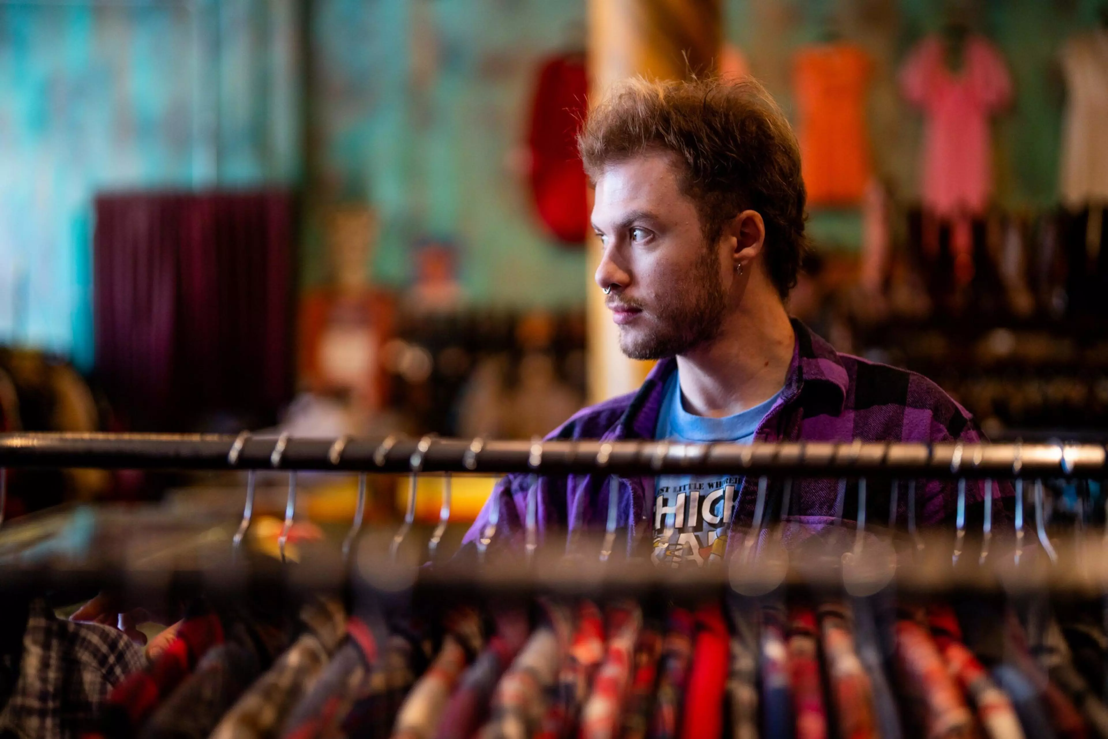 A young man with a beard and mustache looks to the side while standing behind a rack of colorful clothes in a store.