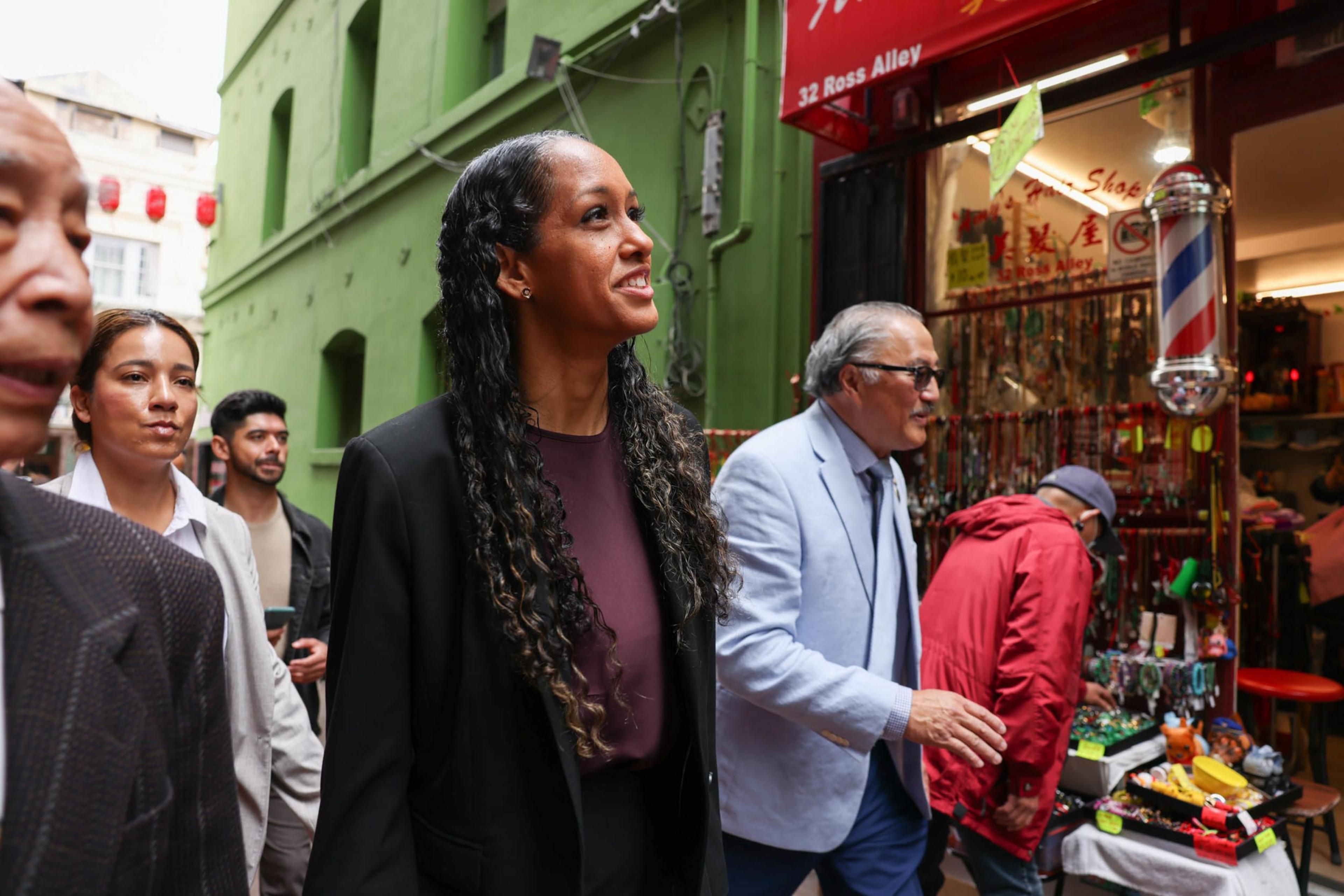 A diverse group of people walk down a narrow street past a colorful shop with a barber pole and various items on display.