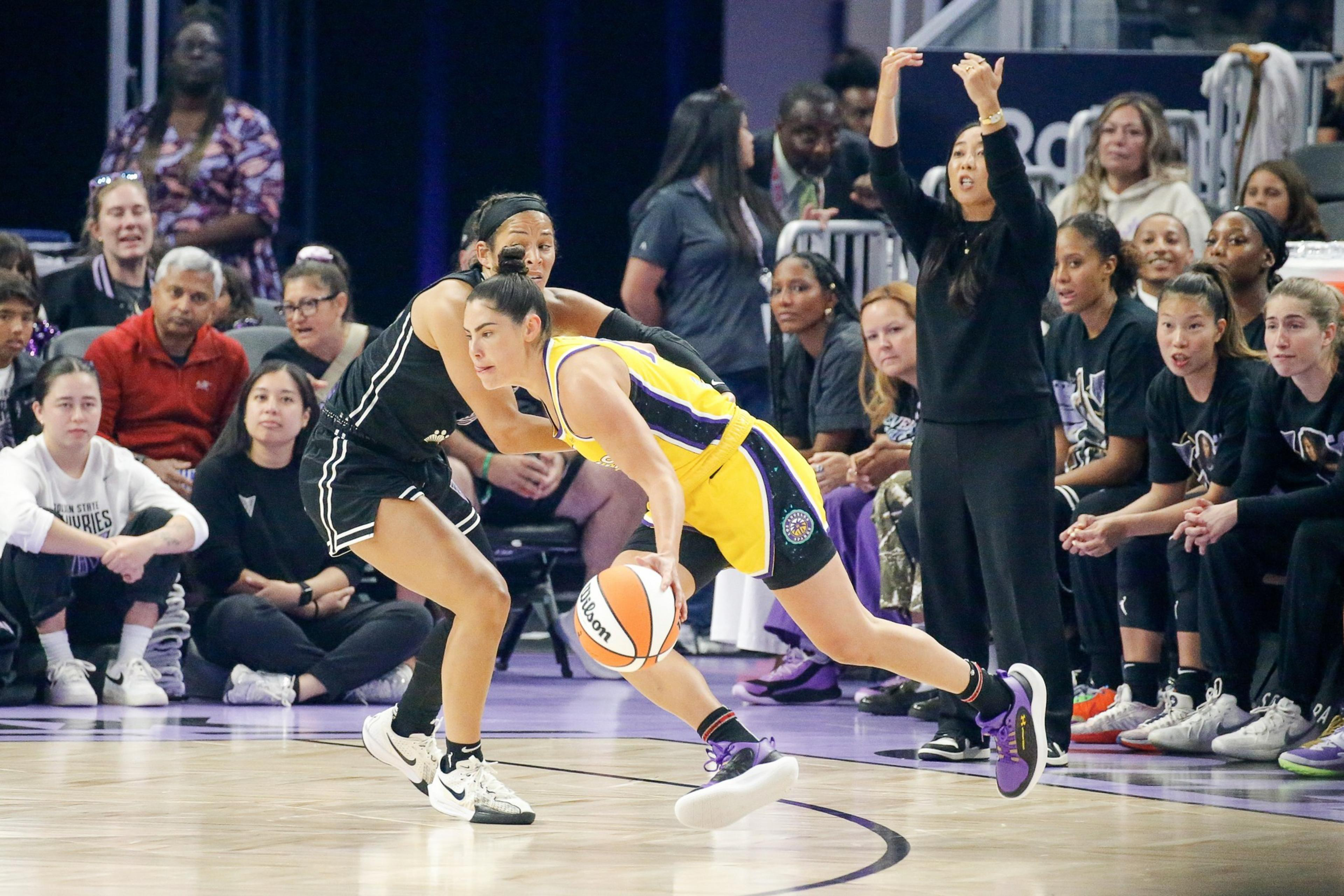 A basketball player in a yellow uniform dribbles past a defender in black, while the crowd and team on the bench intently watch the play unfold.