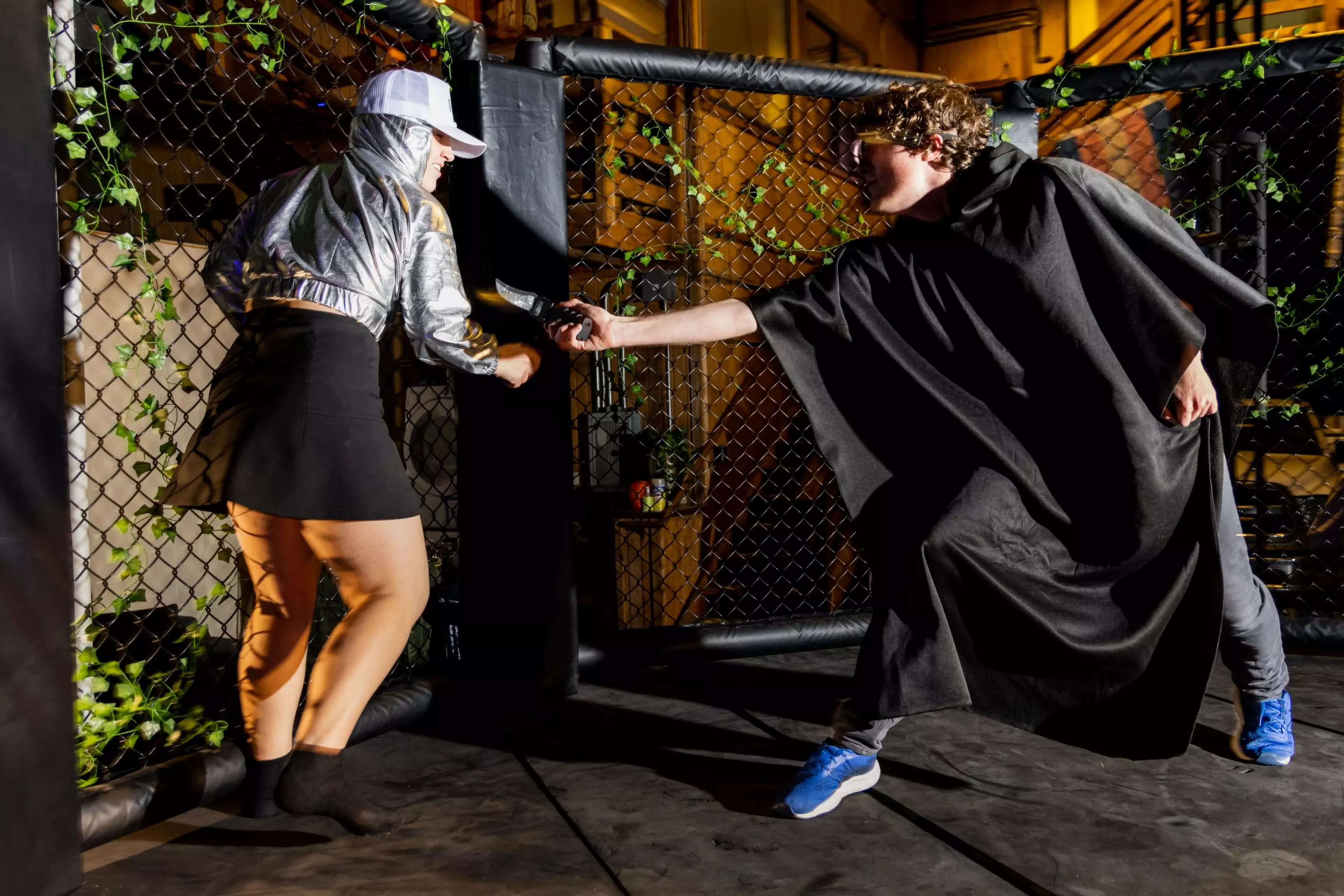 A person in a silver jacket and skirt reaches out while another in a black cloak and blue shoes points a gun toward them inside a fenced area.