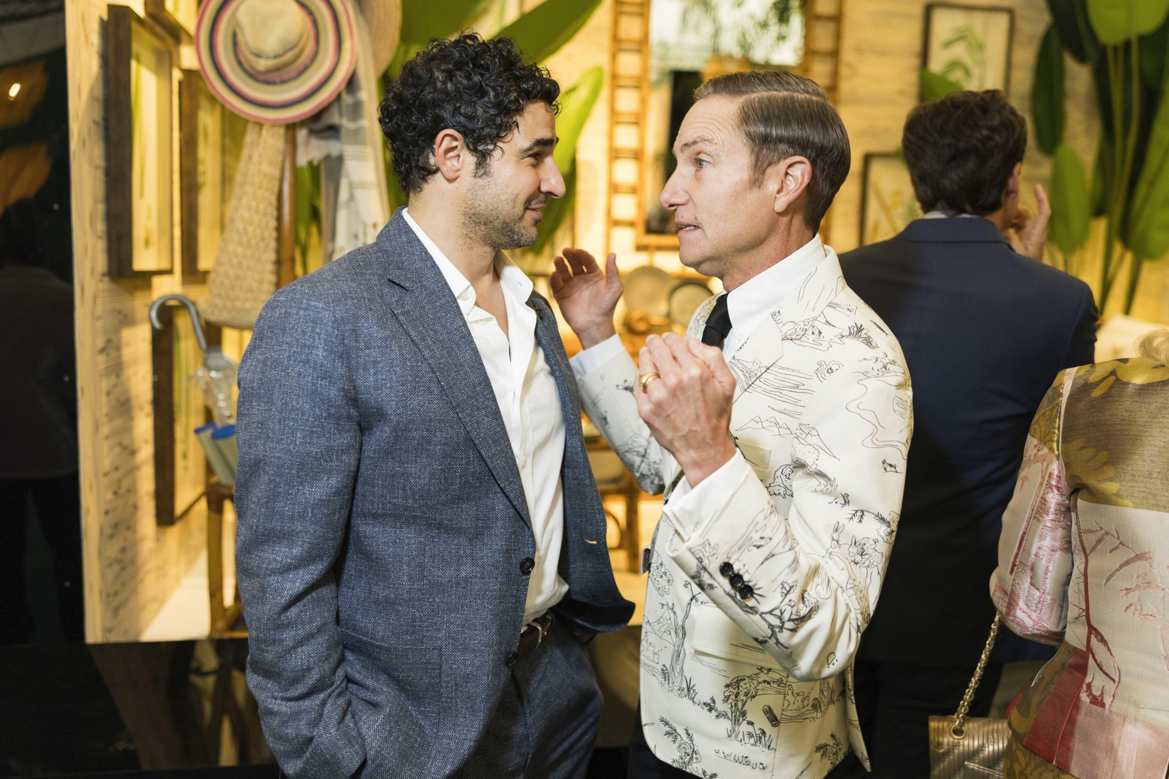 Two men dressed in suits are engaged in a close conversation; one wears a gray suit, the other a patterned white blazer with a black tie.