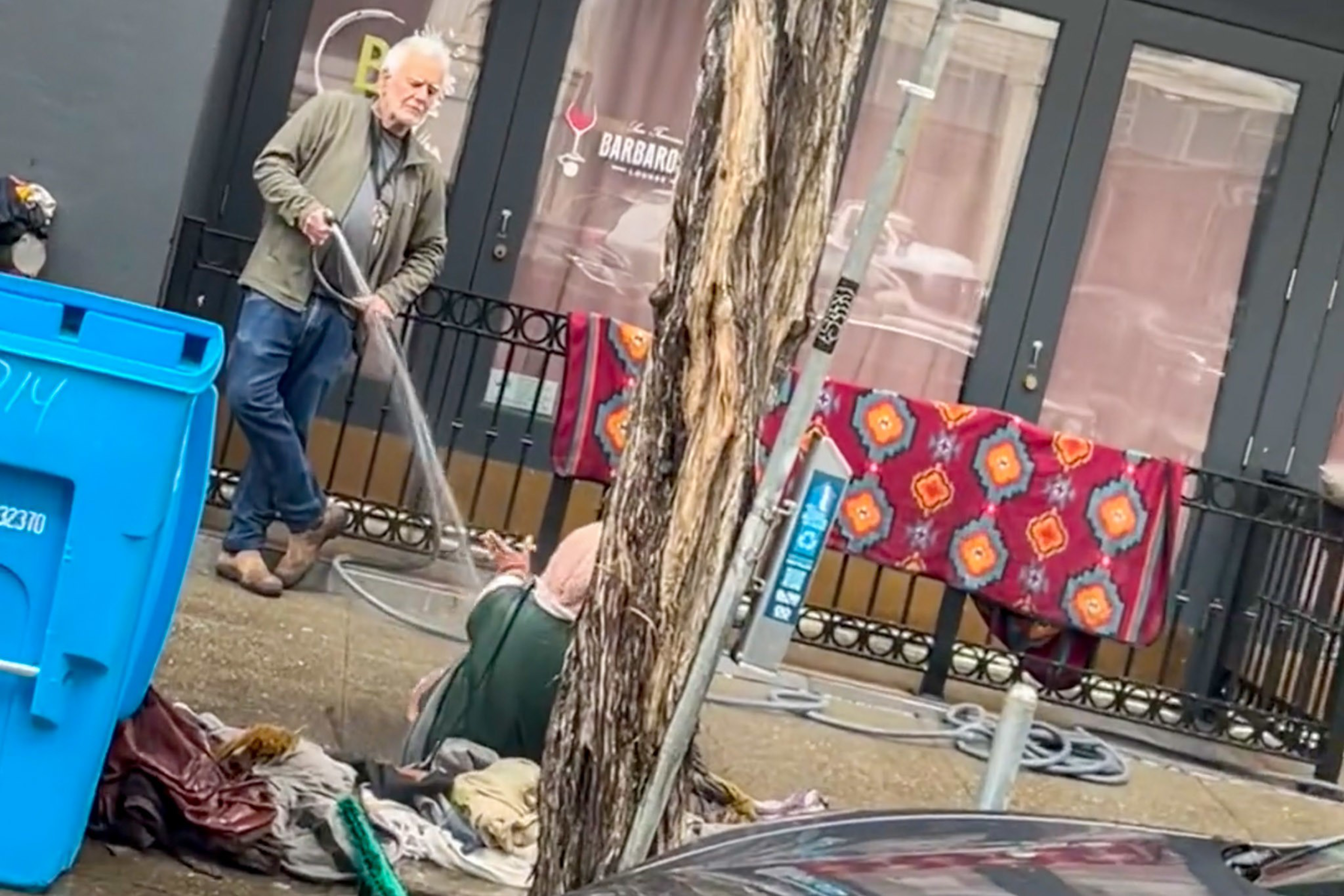 An older man sprays water from a hose at a person sitting on the ground surrounded by clothing and blankets near a storefront with colorful fabric draped on the railing.