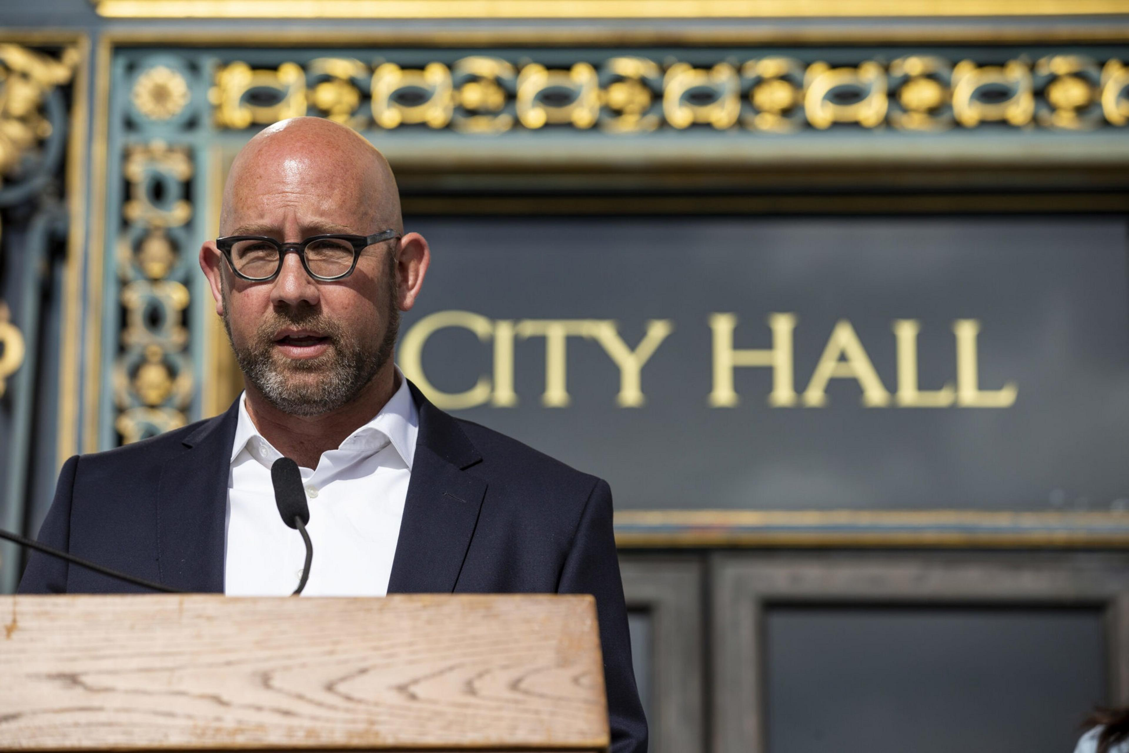 A bald man with glasses speaks at a podium marked â€œCITY HALLâ€ in the background.