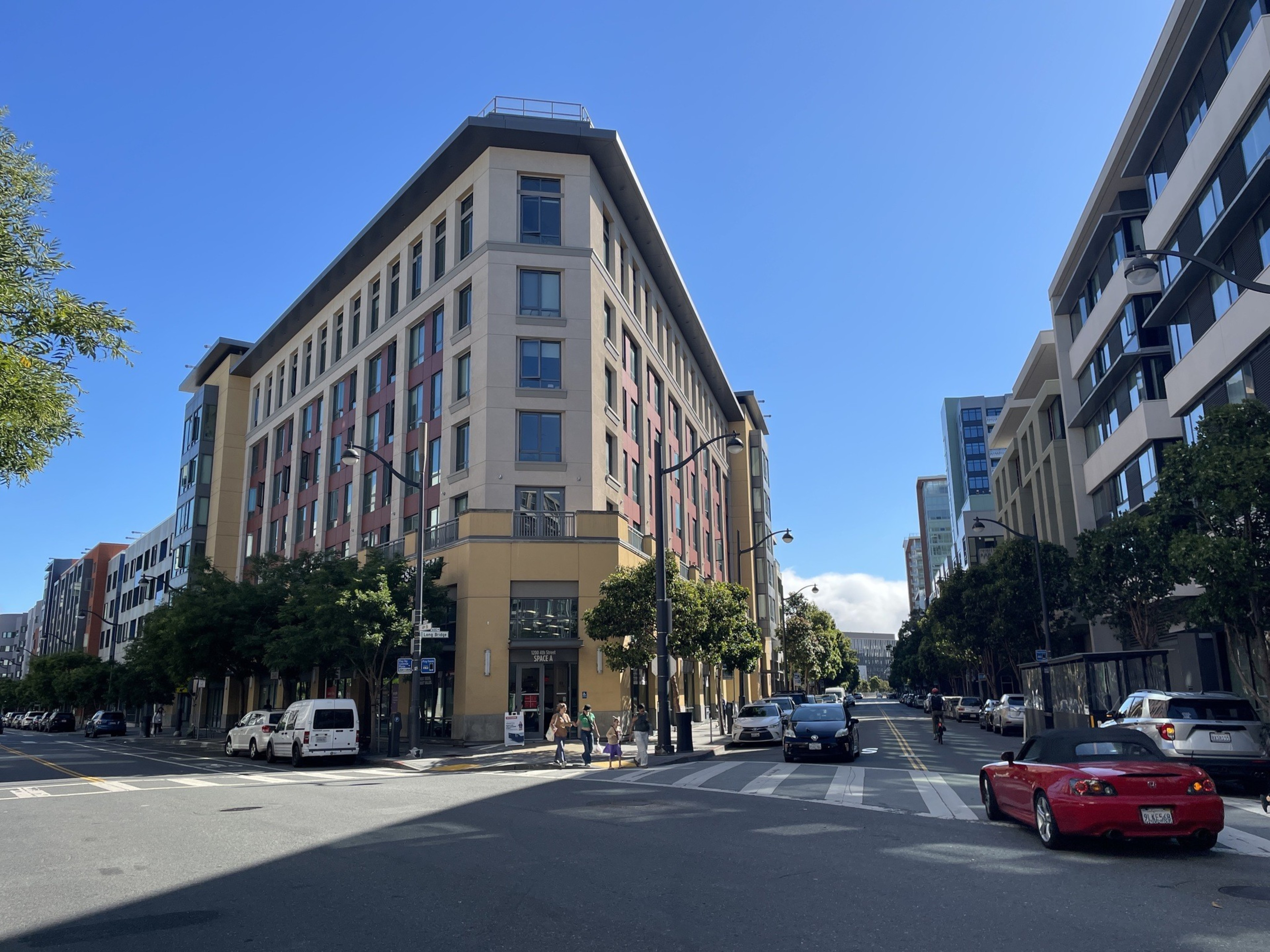 A city street with a six-story beige and red building at the corner, cars parked along the road, trees lining the sidewalks, and clear blue sky above.