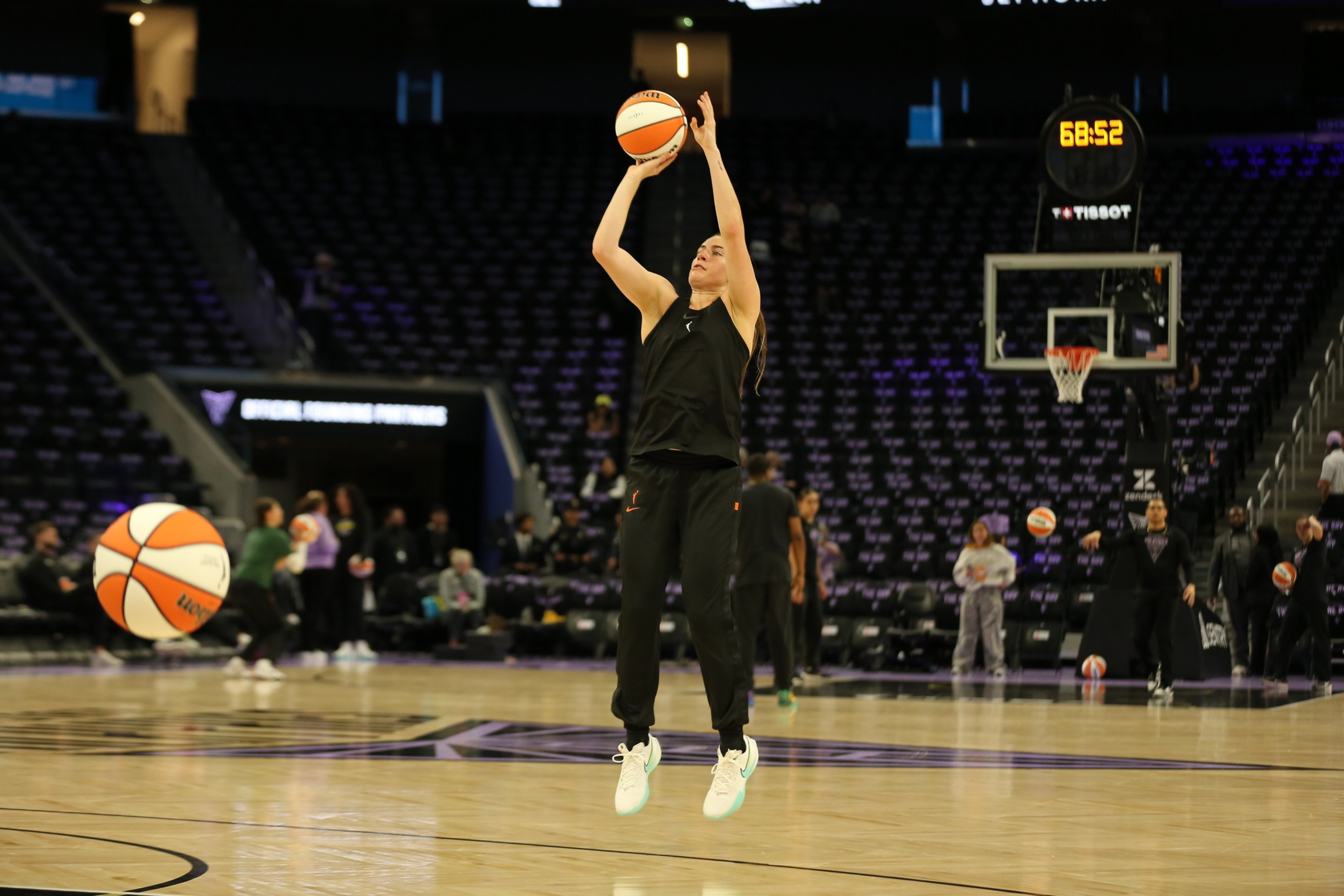 A basketball player in black athletic wear jumps to shoot a basketball on an empty court with several players and basketballs in the background.