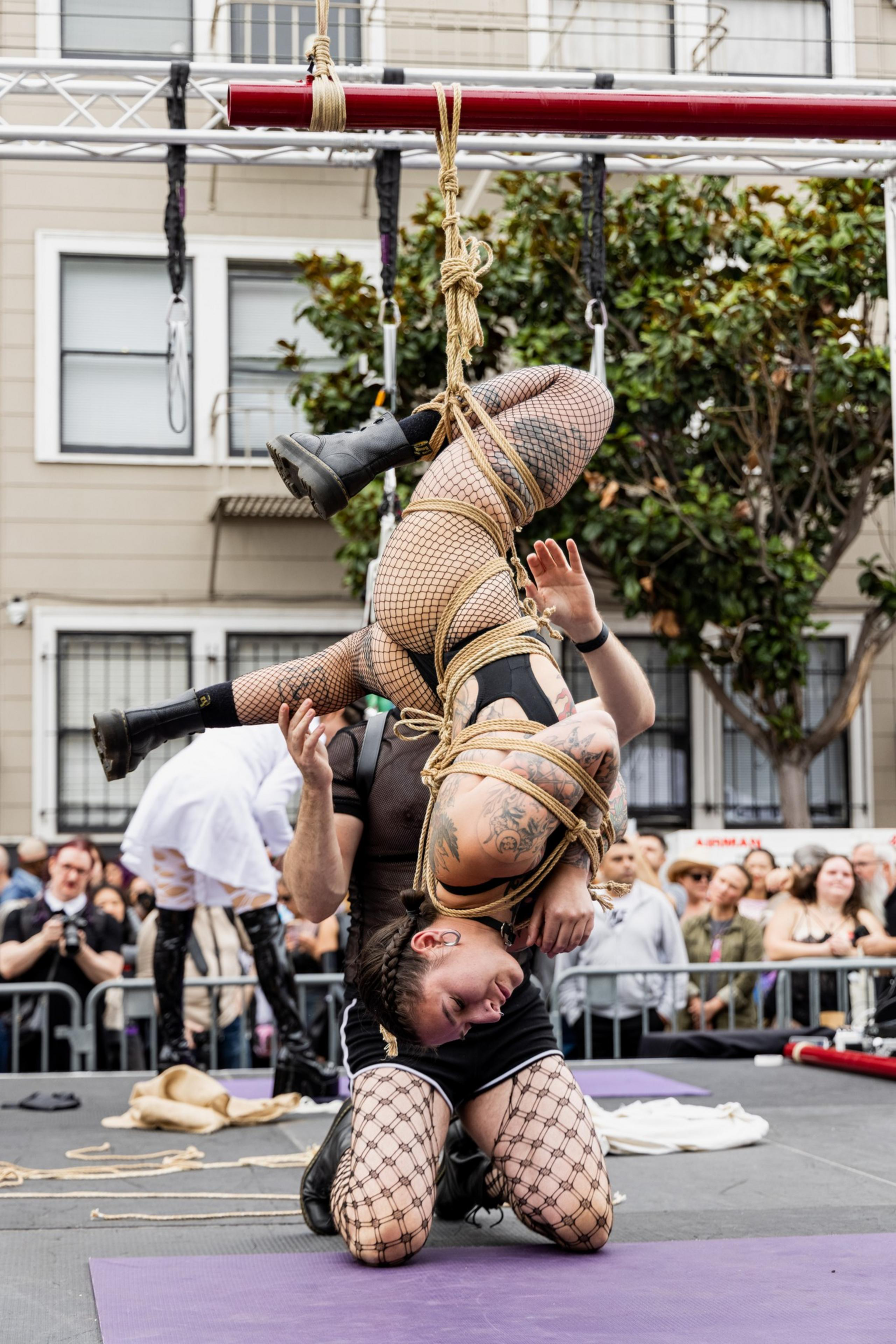 A person in fishnet stockings and boots is suspended upside down by intricate rope bondage, supported by another kneeling person, with a crowd watching.