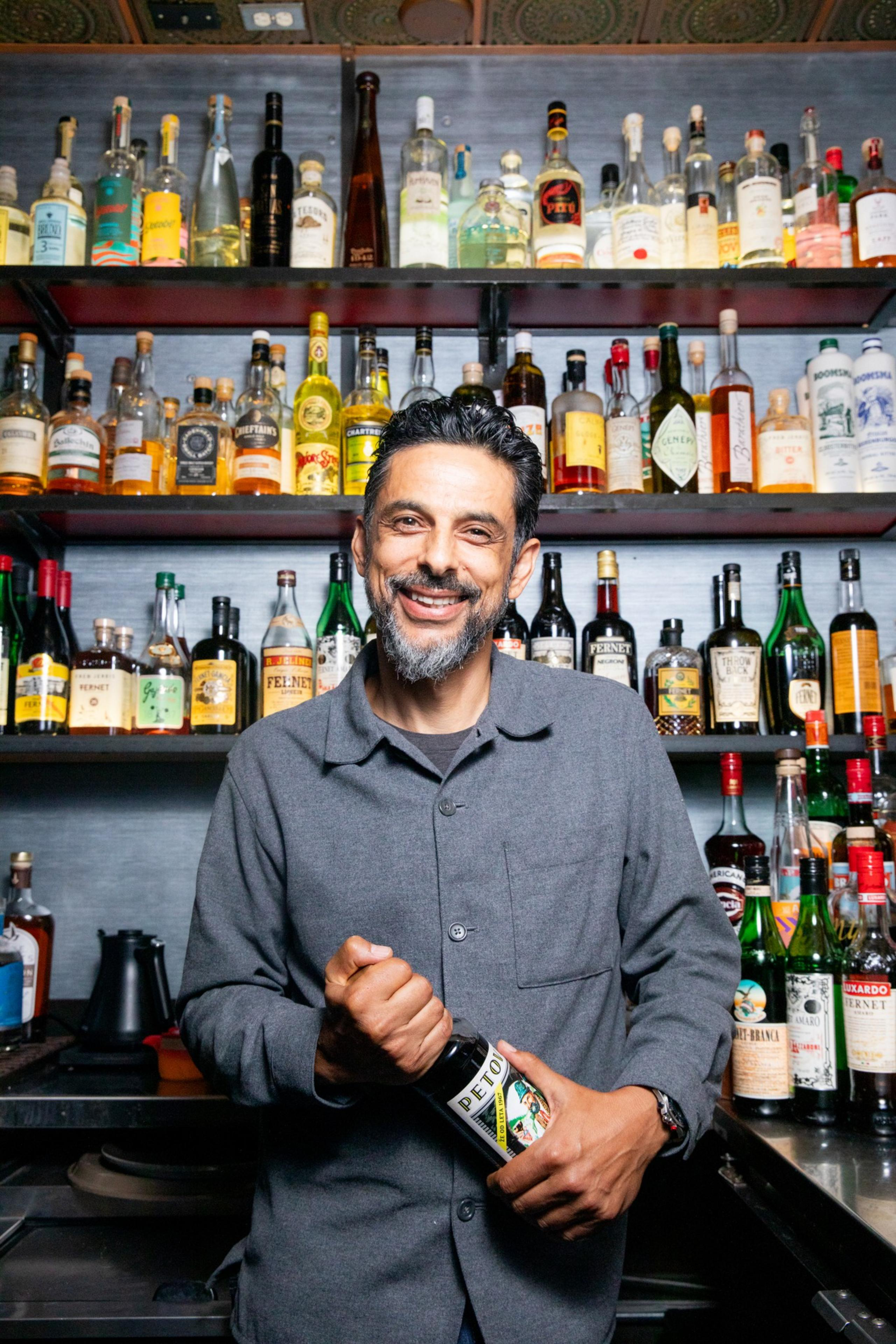 A smiling man in a gray shirt stands in front of shelves filled with various colorful liquor bottles. He is holding a bottle labeled &quot;PETONI&quot; in his hands.