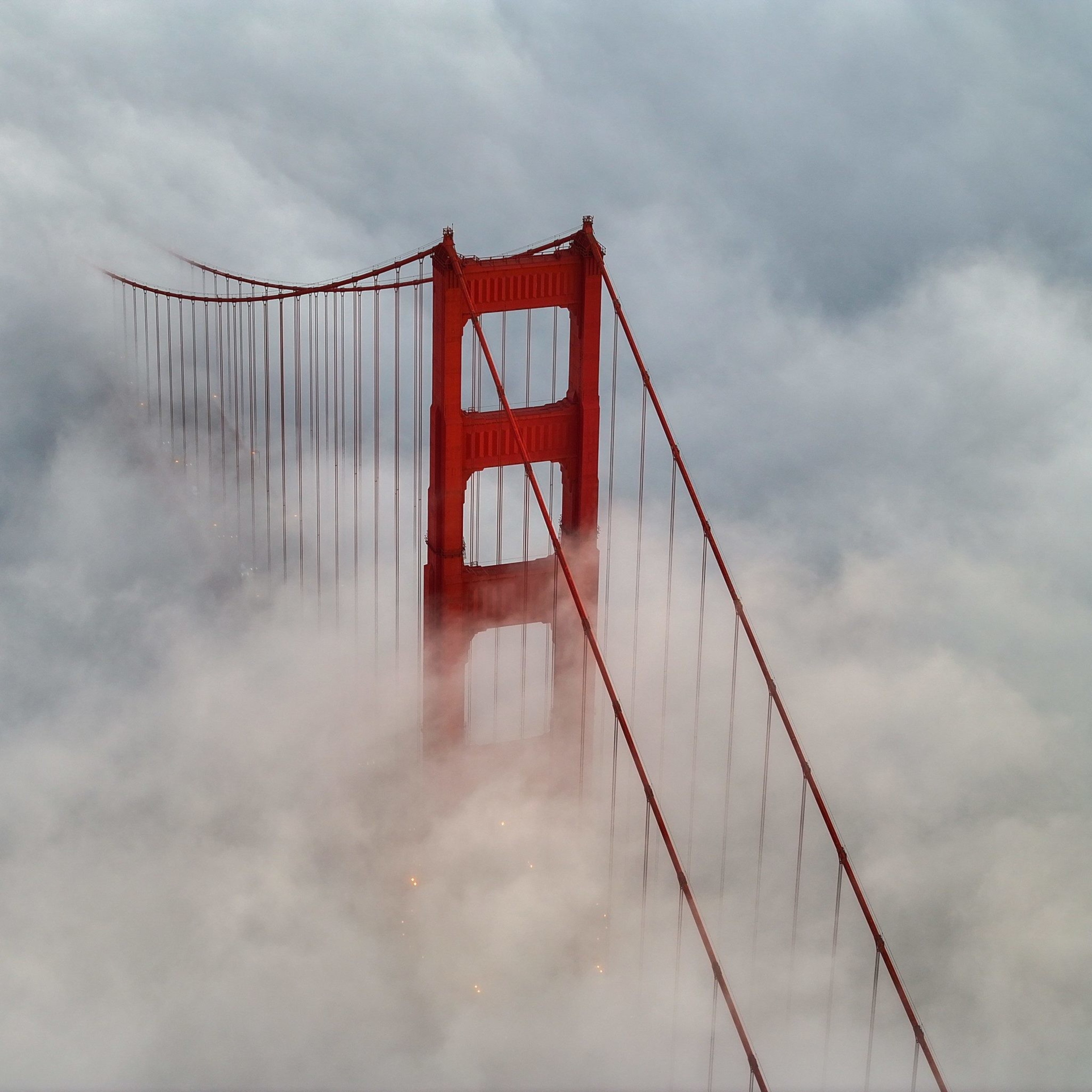 A tall, red suspension bridge partially shrouded in thick clouds, with its vertical towers and cables peeking through, creating an ethereal, misty scene.