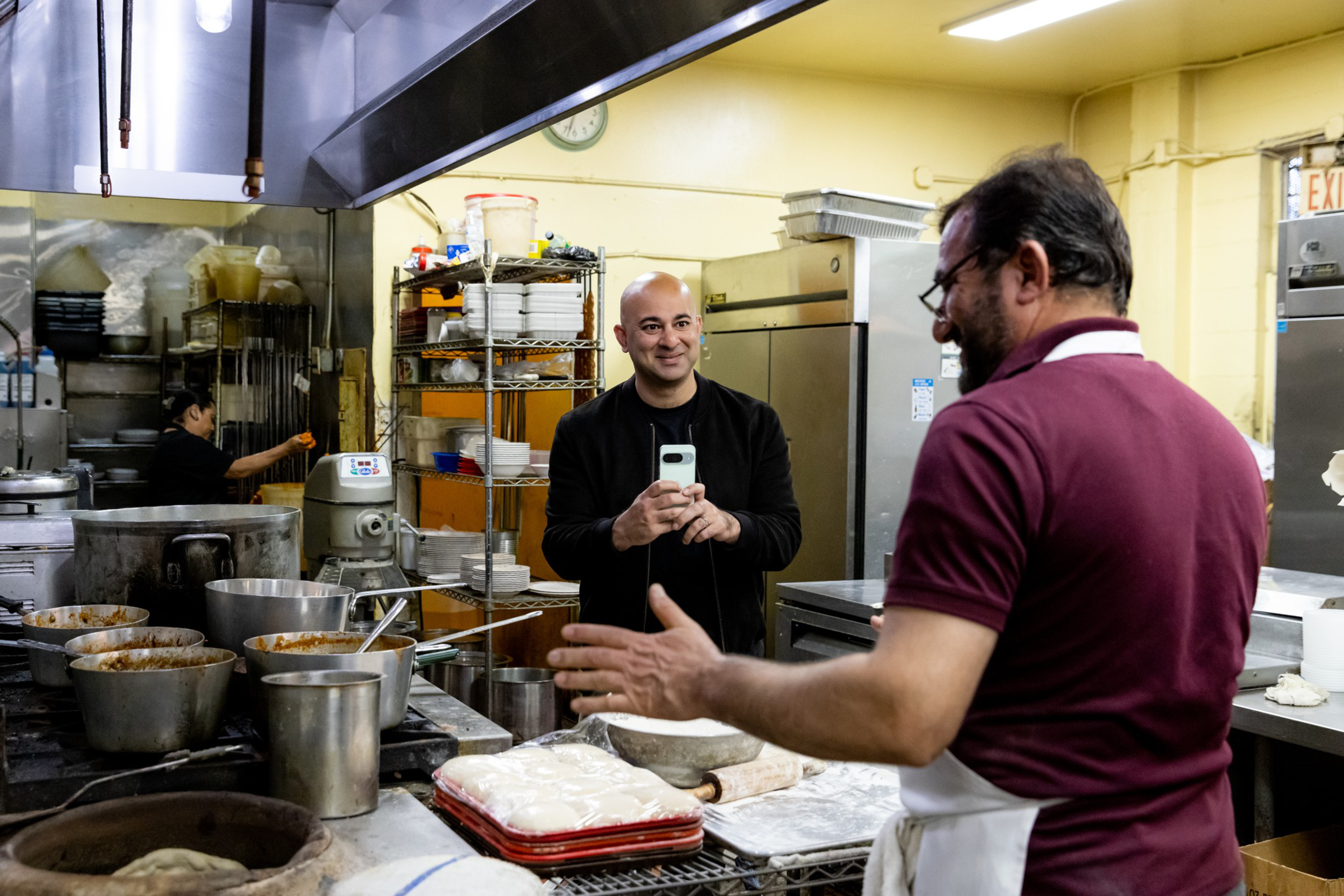 A man in a maroon shirt and apron gestures while talking to a smiling man holding a phone, surrounded by cooking pots and dough in a kitchen.