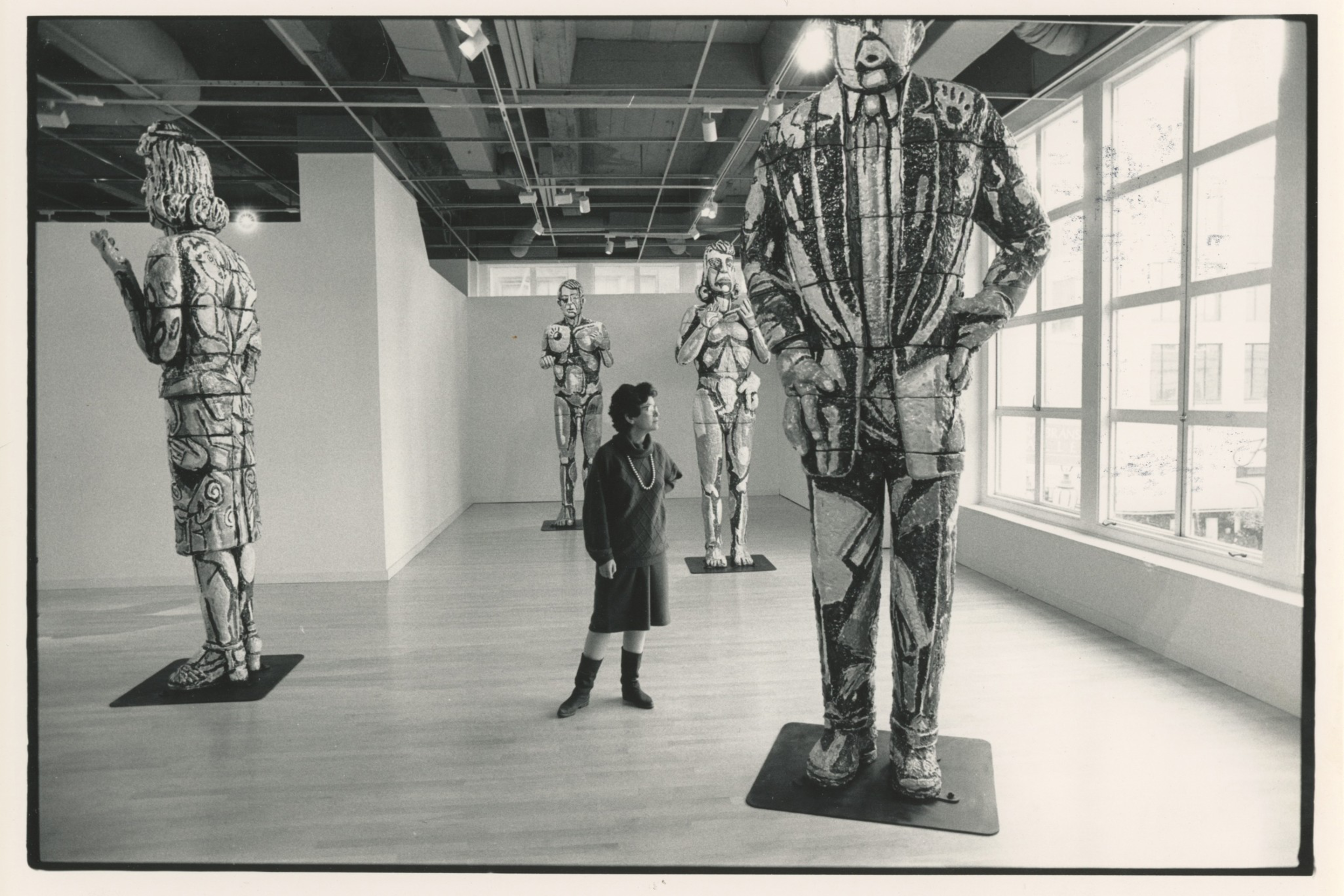 A woman stands in a gallery surrounded by five large, textured sculptures of human figures in various poses, near large windows letting in natural light.