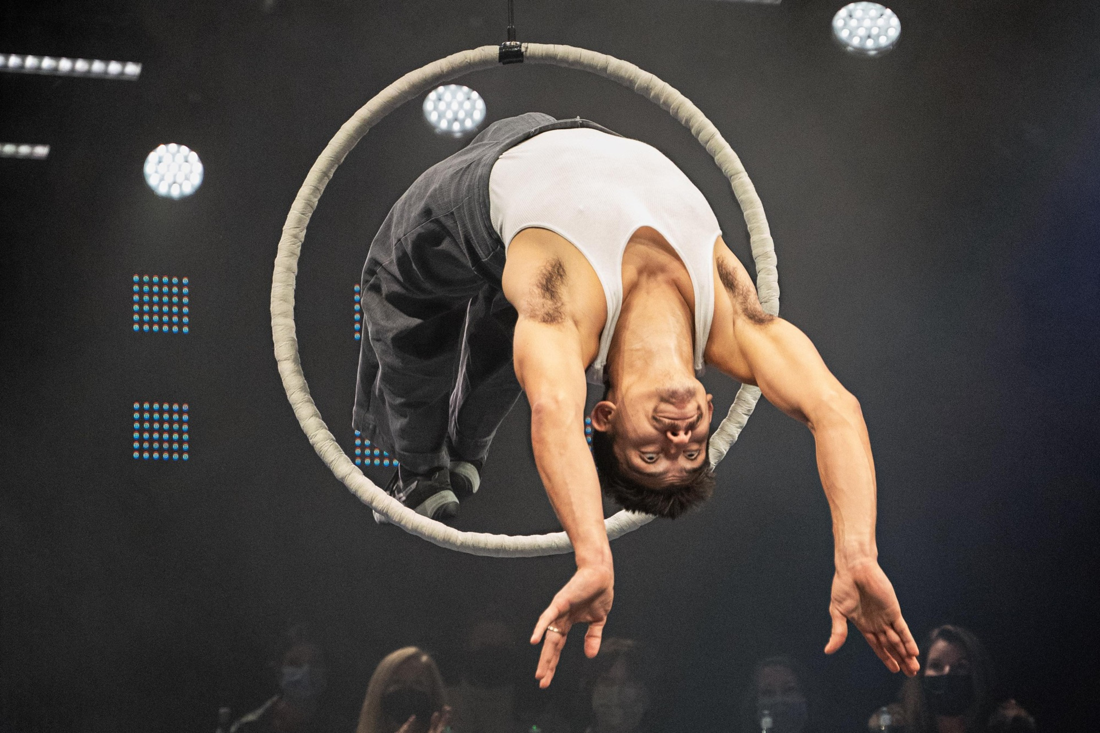 A performer in a white tank top and dark pants is upside down, balancing in a hoop, suspended with stage lights above and an audience below.