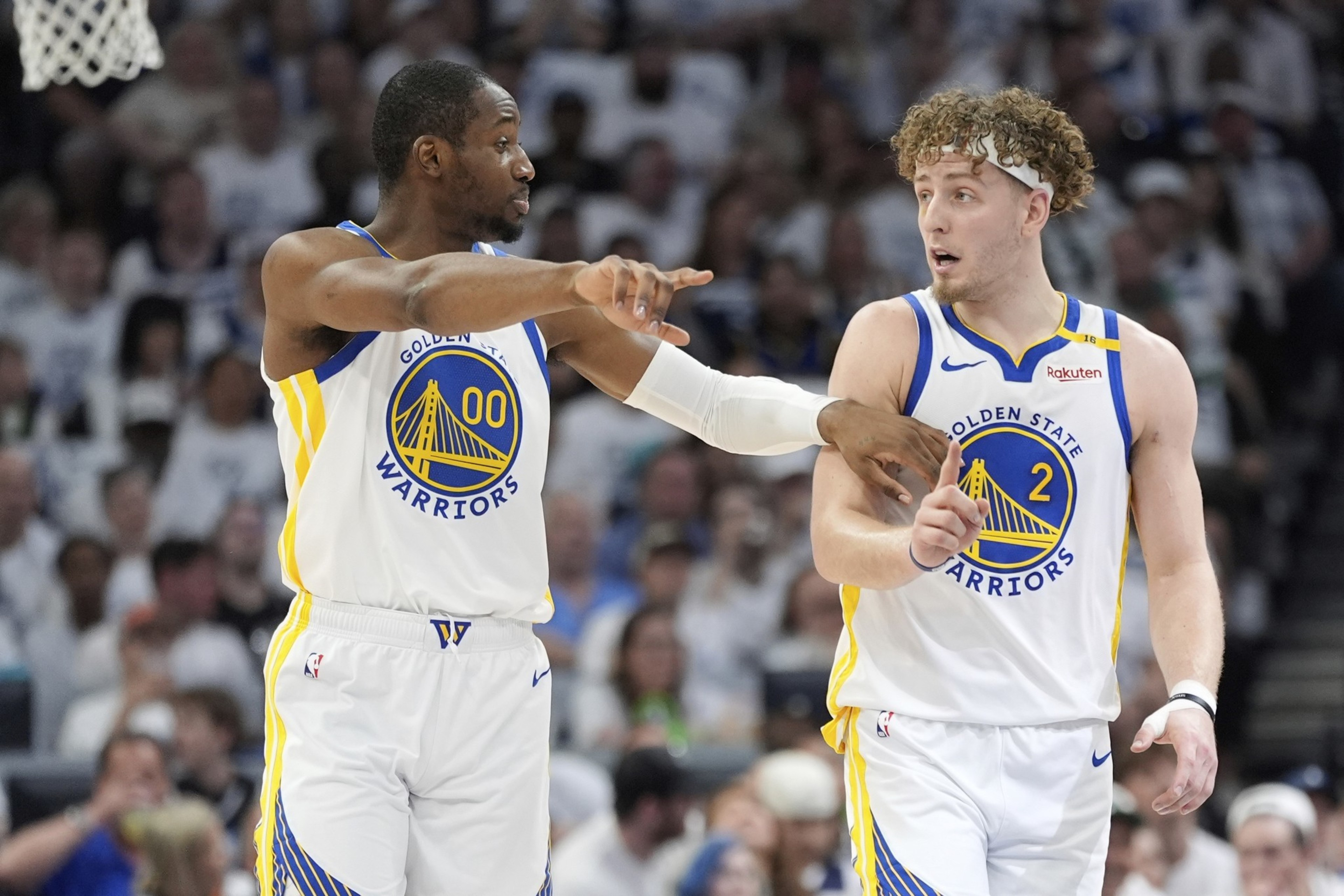 Two basketball players in Golden State Warriors jerseys are on the court during a game. One player points while the other looks ahead with an attentive expression.
