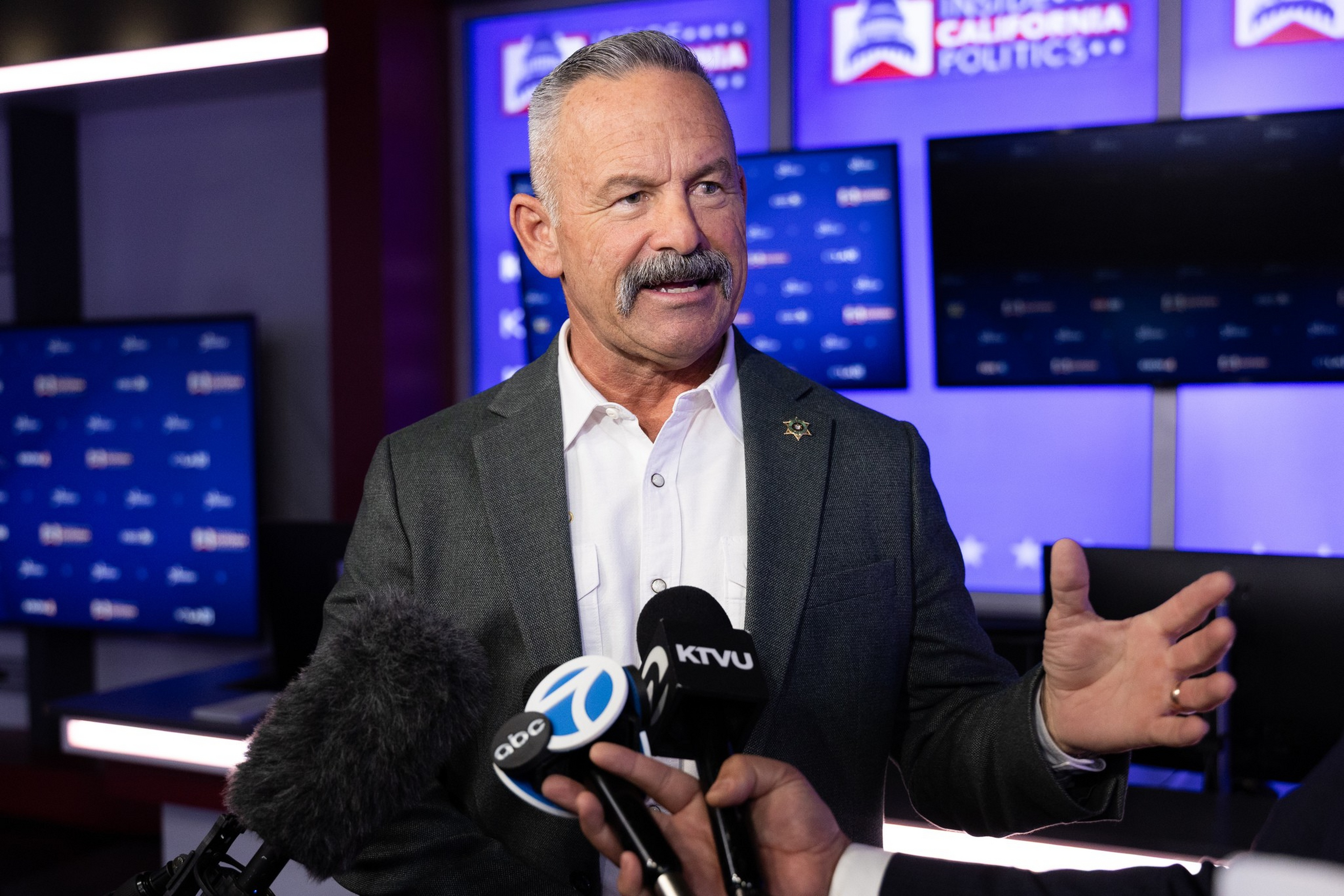 A man with gray hair and a mustache speaks while gesturing, facing multiple microphones labeled abc 7 and KTVU in a political studio setting.
