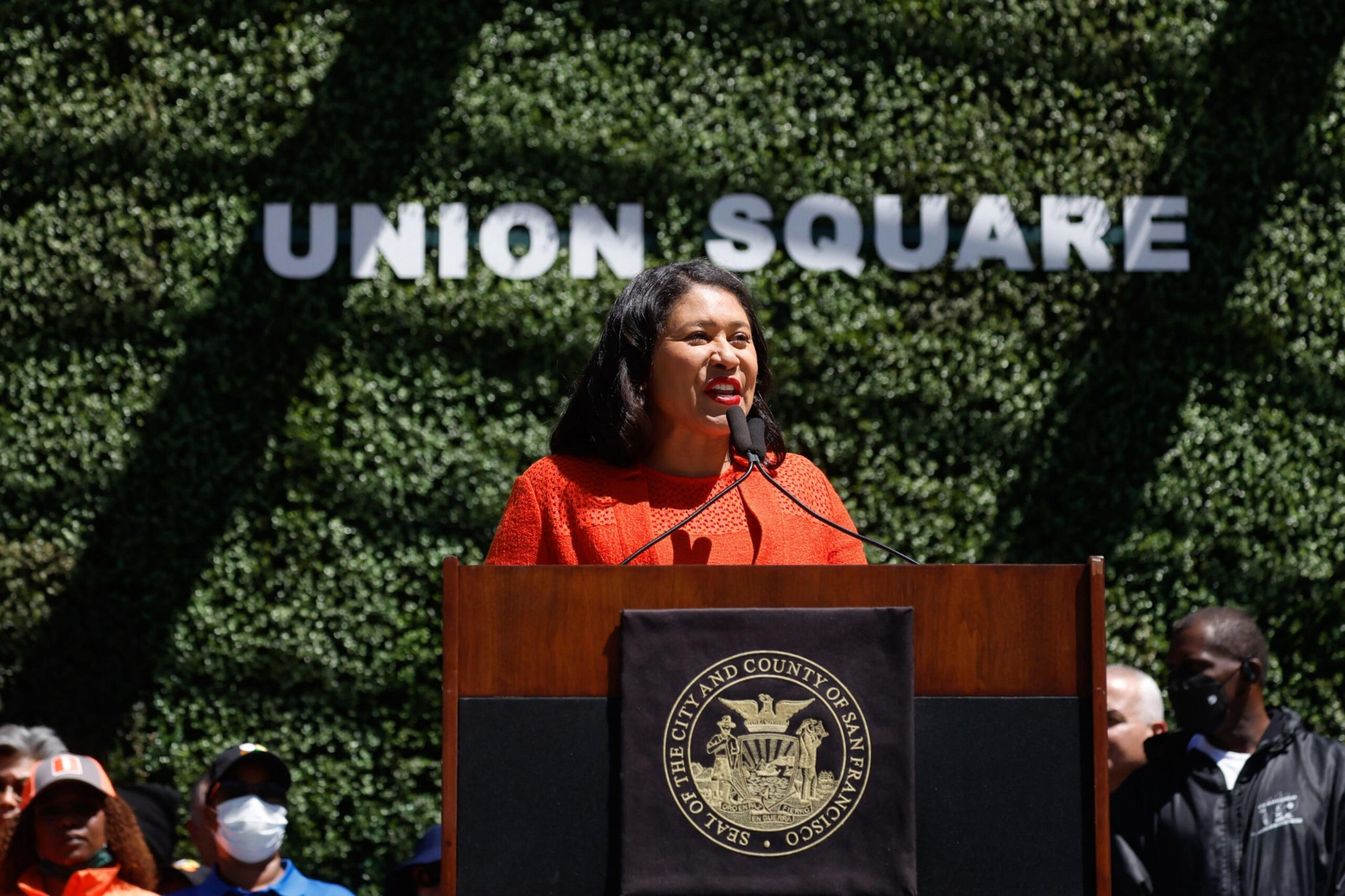 A woman in an orange outfit speaks at a podium with the San Francisco city seal, in front of a green wall with "UNION SQUARE" letters.