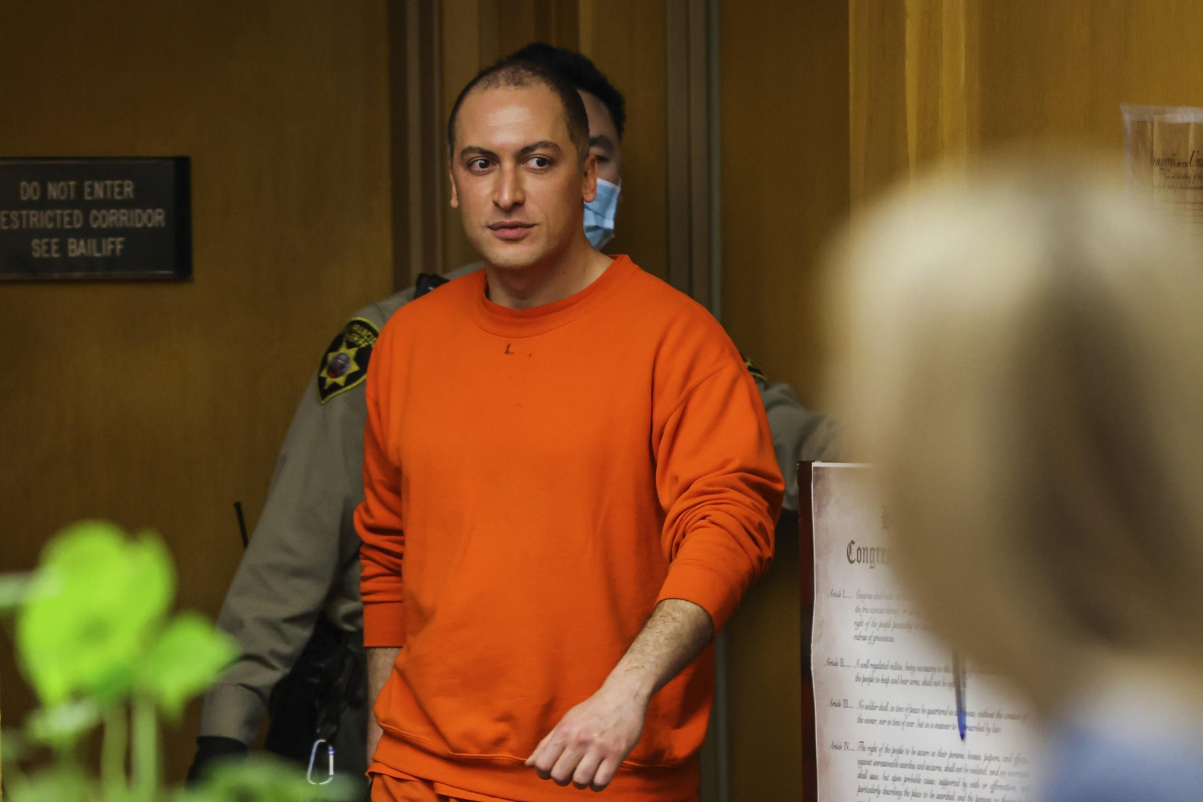 A person in an orange jumpsuit walks in a hallway, accompanied by a law enforcement officer. Papers and a sign are visible on a wall in the background.