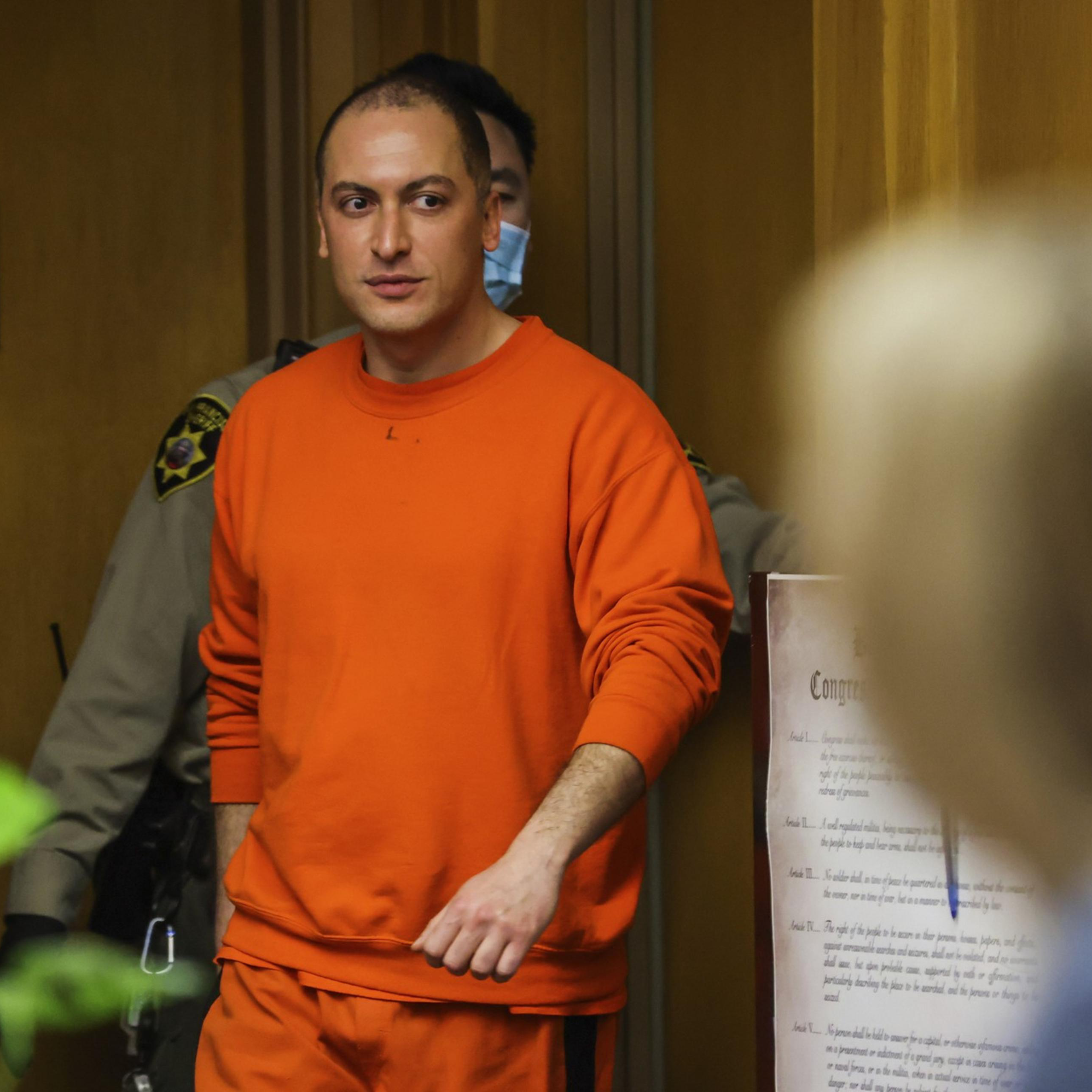 A person in an orange jumpsuit walks in a hallway, accompanied by a law enforcement officer. Papers and a sign are visible on a wall in the background.