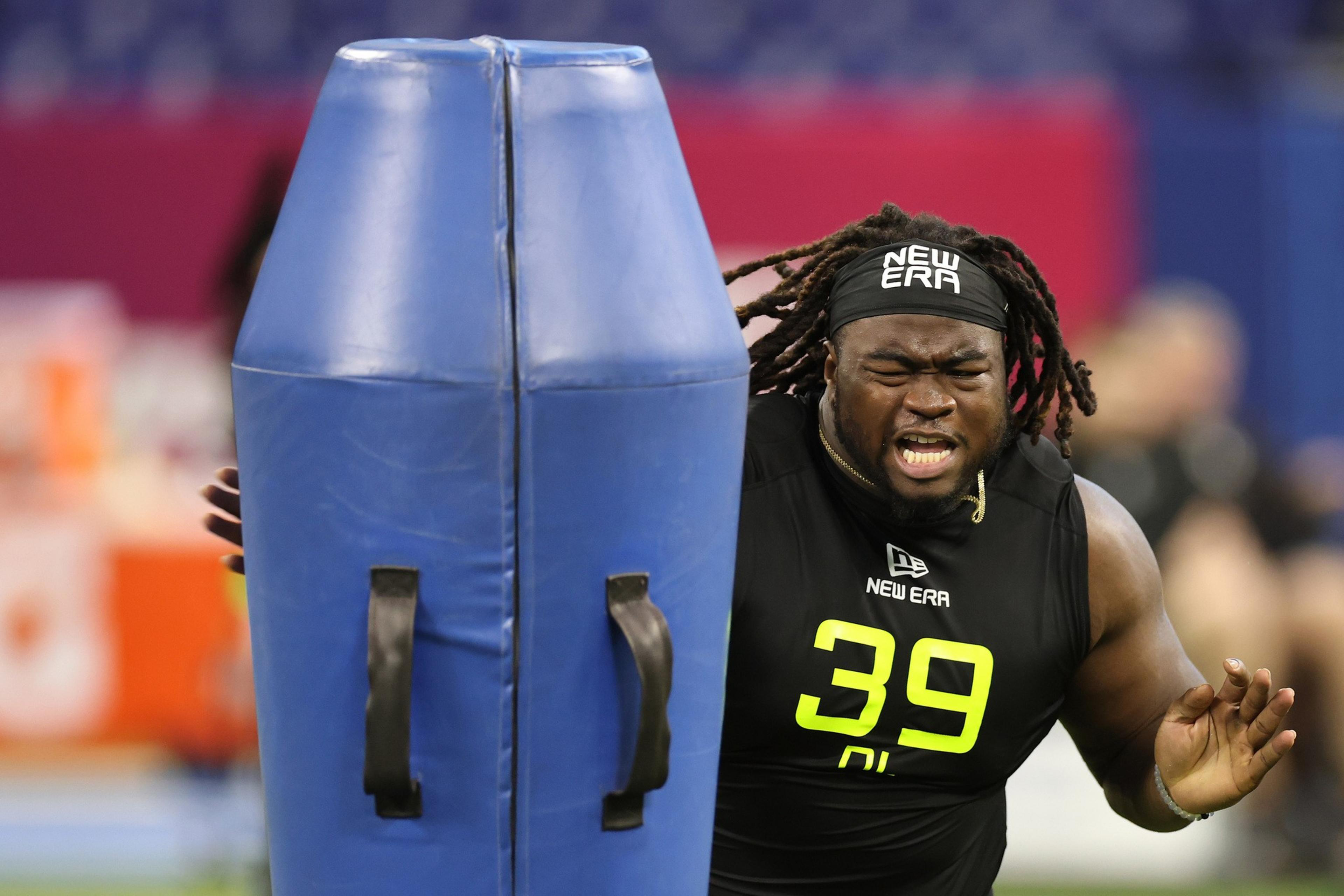 A man wearing a black sports shirt with "39" on it pushes against a large blue padded obstacle. He appears to be exerting force, with a focused expression.