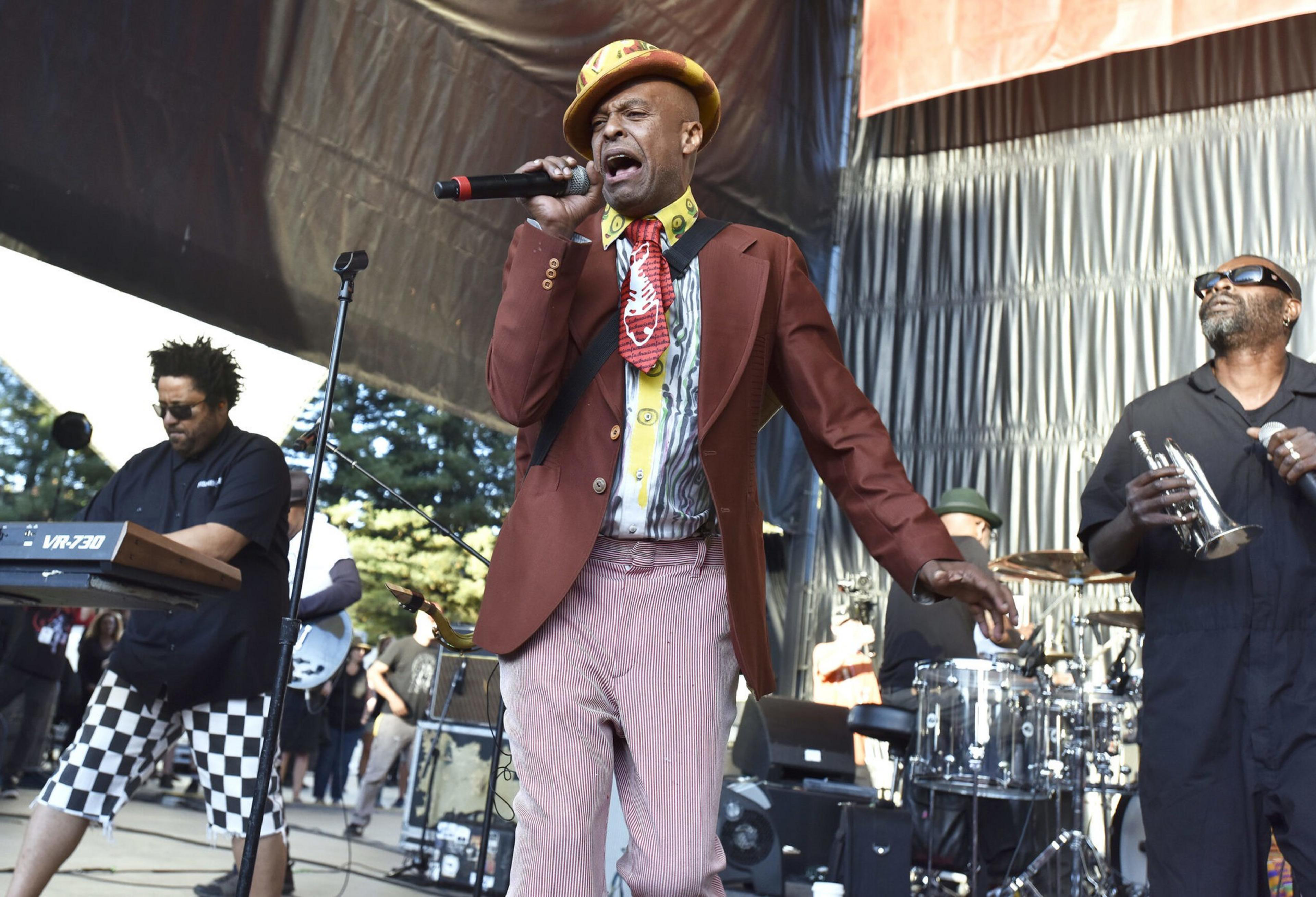 A singer in a colorful outfit passionately performs with a band, including a keyboardist in checkered shorts and a trumpeter wearing sunglasses.