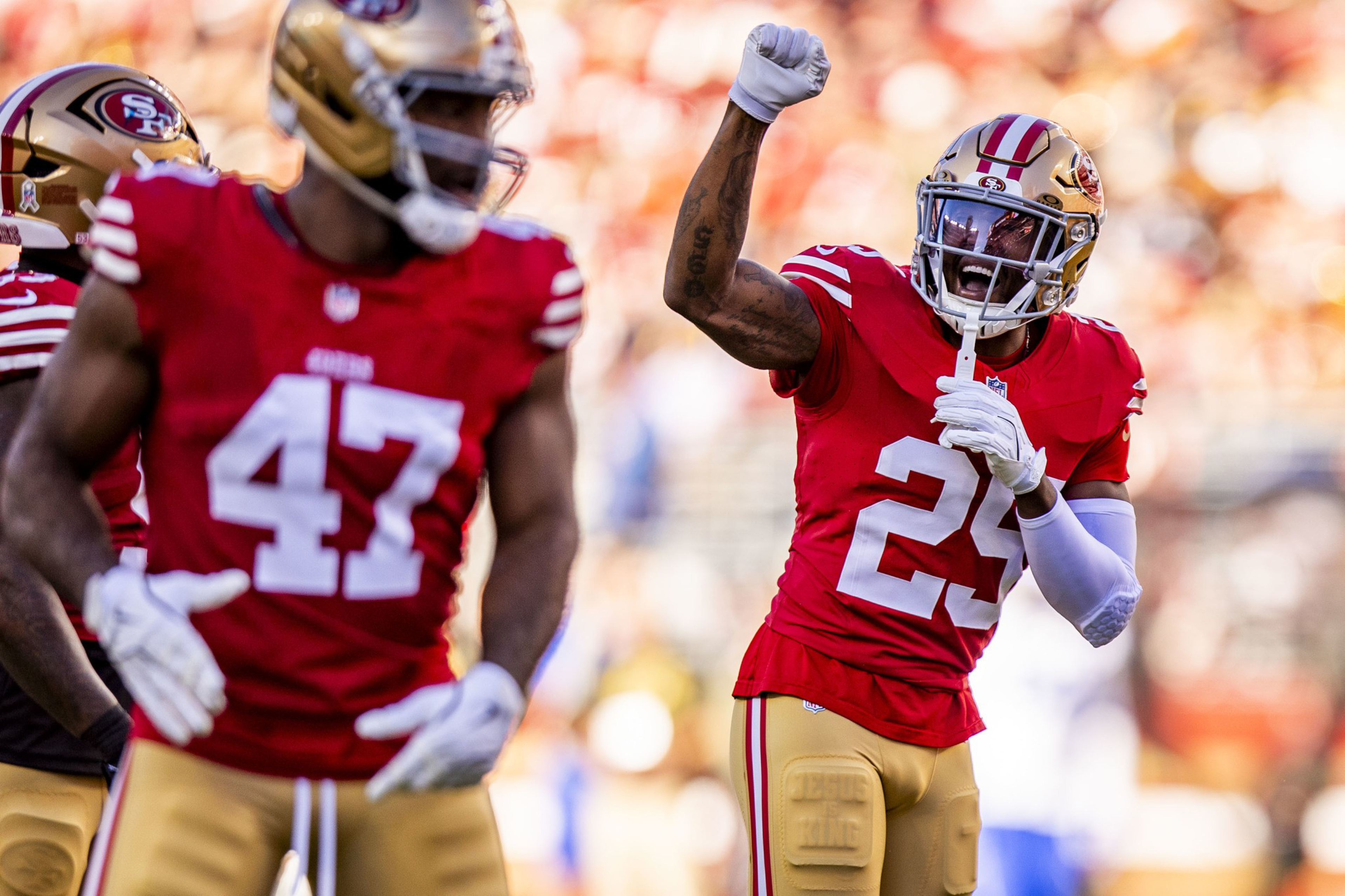 Two San Francisco 49ers players in red jerseys and gold helmets celebrate on the field, with one raising his fist excitedly.