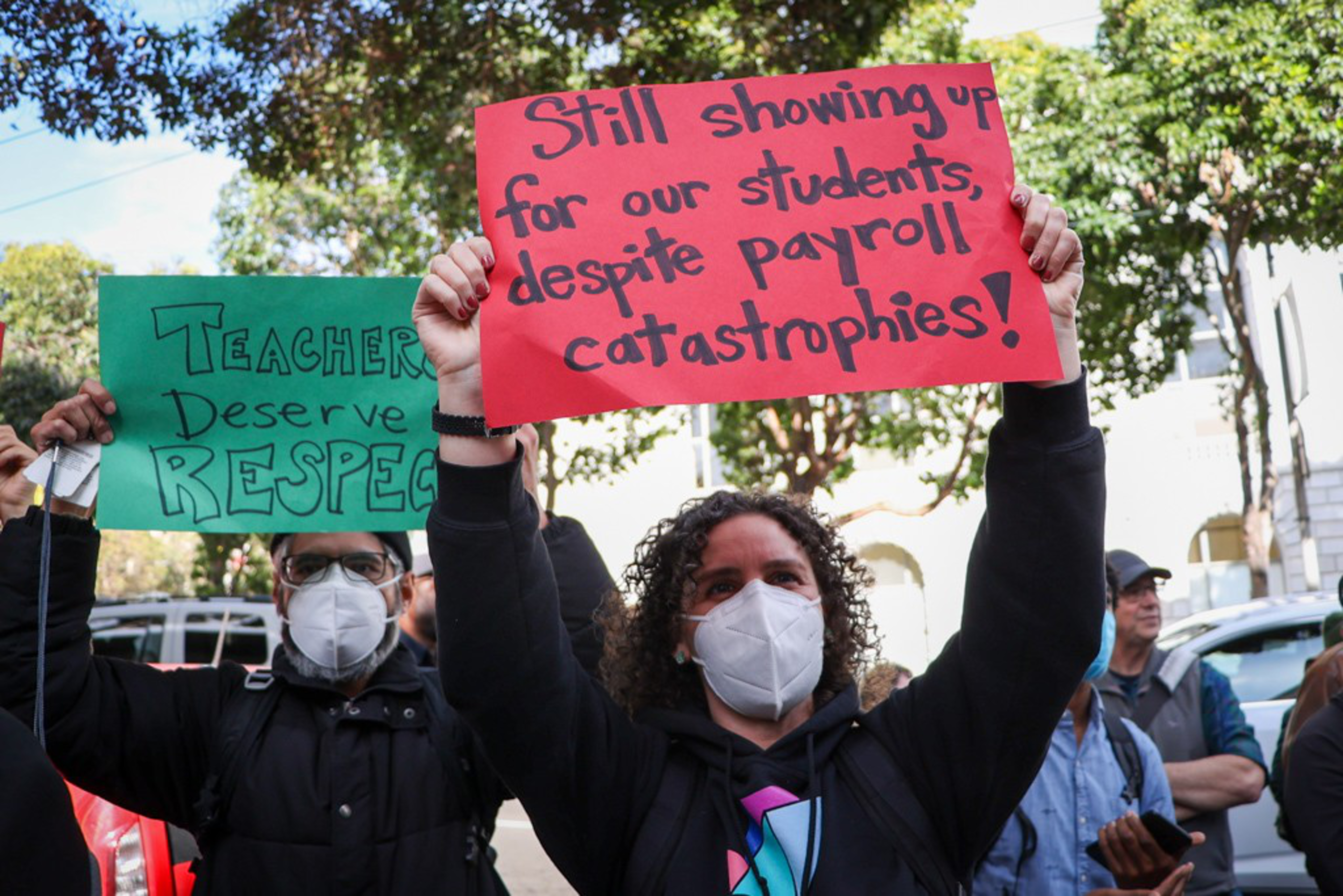 A person wearing a mask holds a red sign reading, "Still showing up for our students despite payroll catastrophes!" while another holds a green sign saying, "Teachers Deserve Respect."