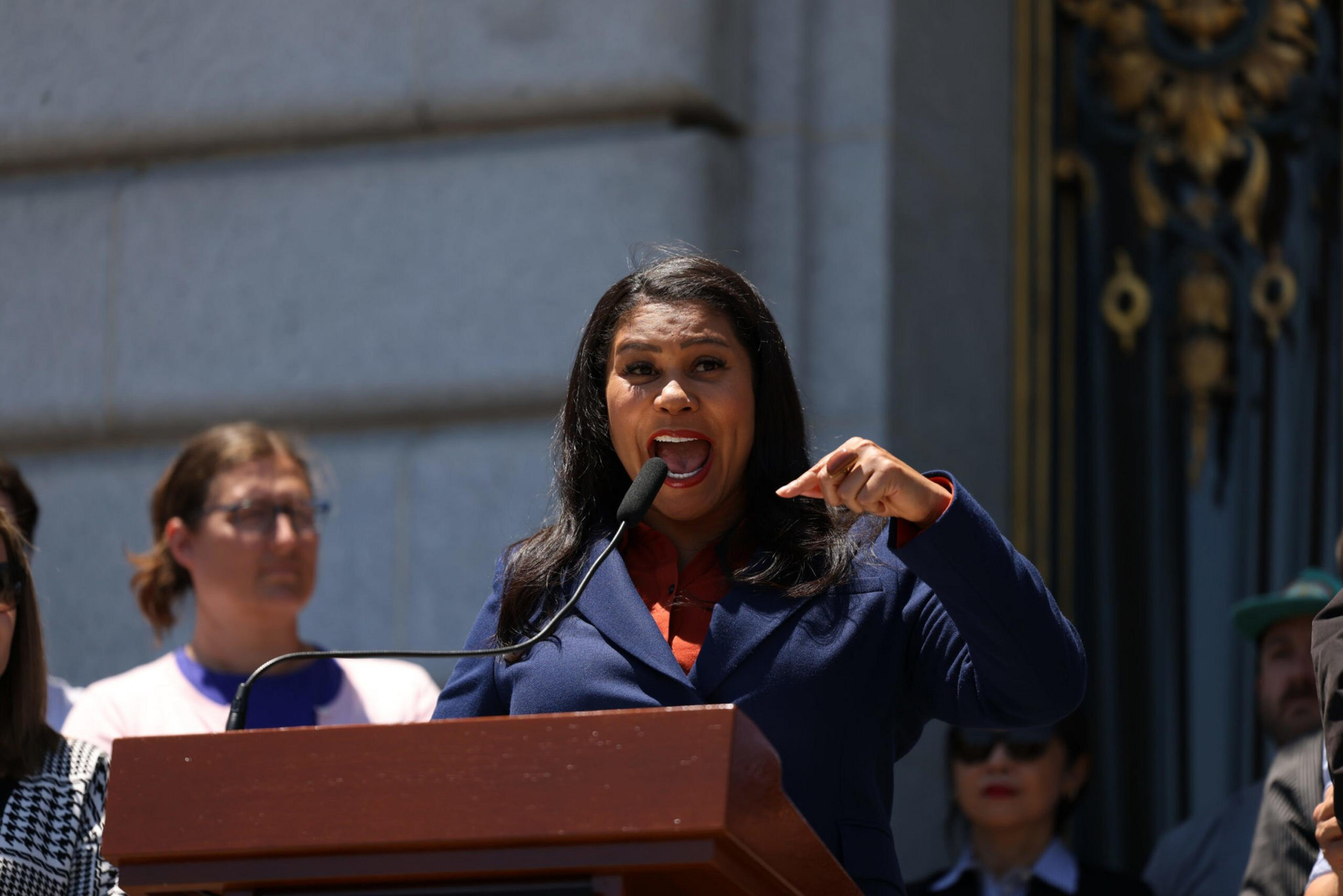 A woman in a blue blazer passionately speaks into a microphone at a podium, surrounded by several attentive people in the background.