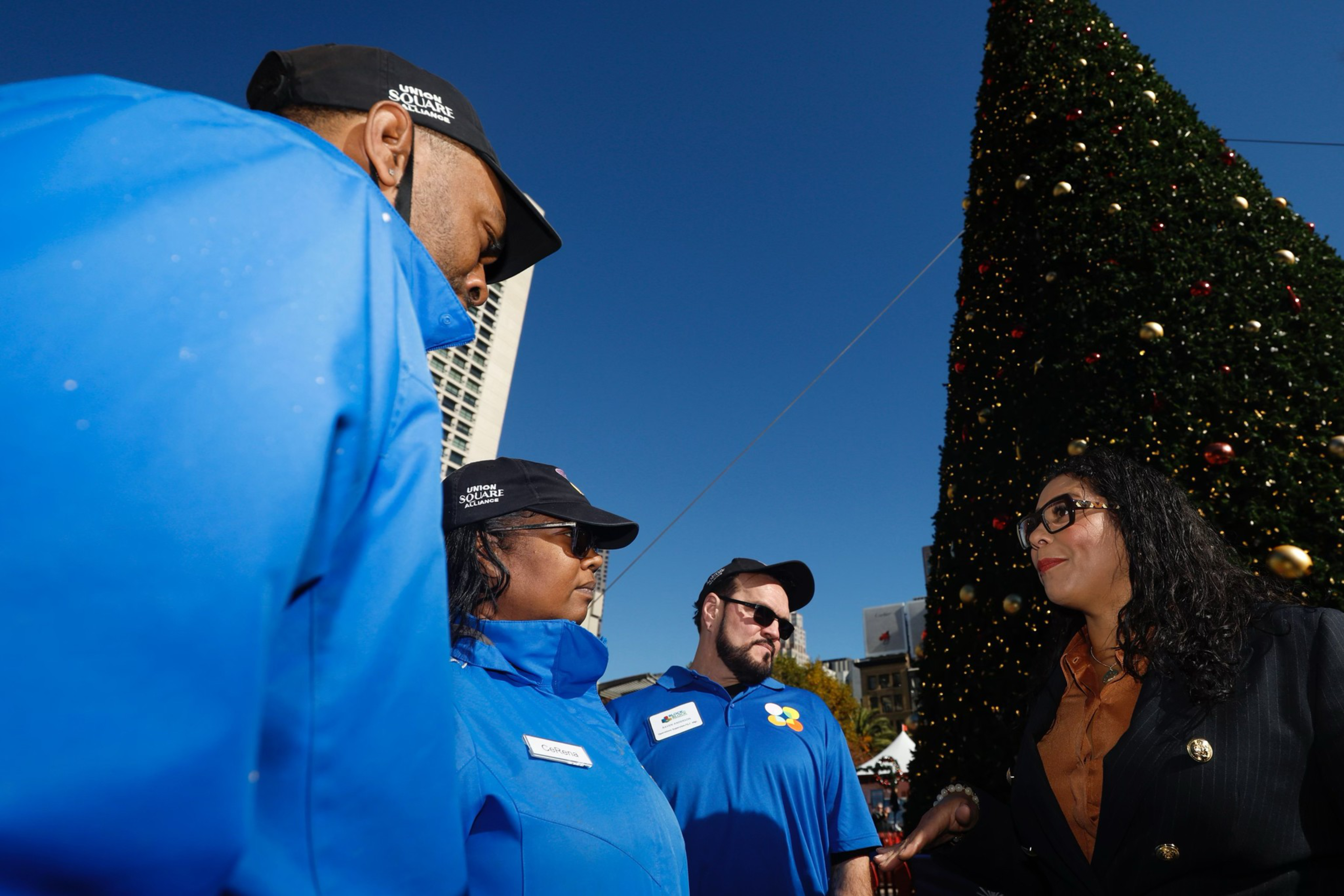 Three people in blue uniforms with name tags and caps talk to a woman in a dark coat near a large decorated Christmas tree outdoors.