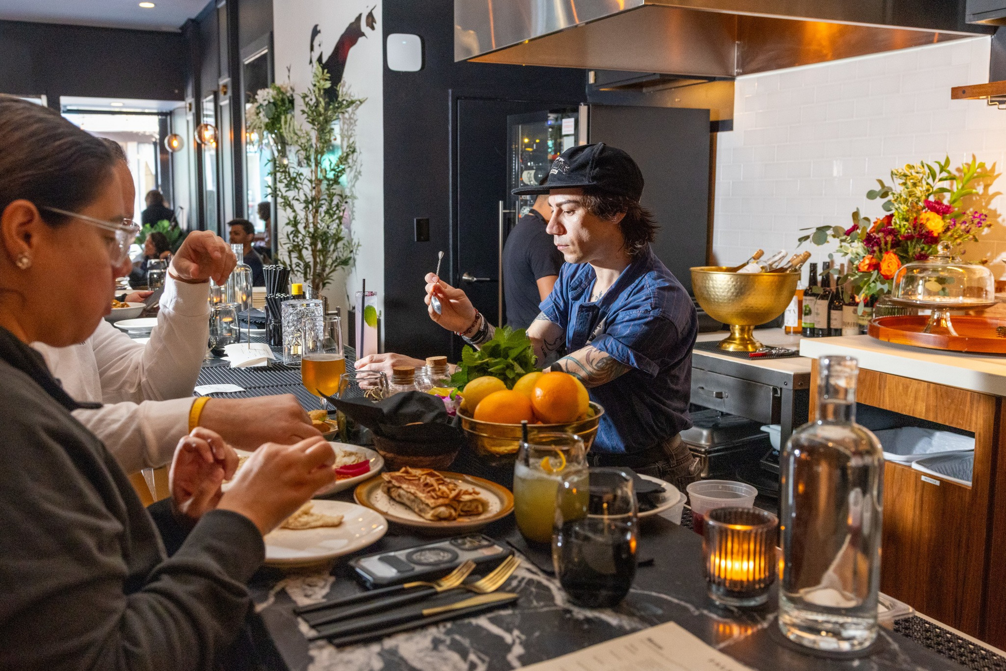 Ένας άντρας με καπέλο και μπλε πουκάμισο ετοιμάζει ένα ποτό πίσω από έναν πάγκο μπαρ ενώ δύο άτομα τρώνε κρέπες στο μπαρ σε ένα ζεστό, μοντέρνο εστιατόριο.