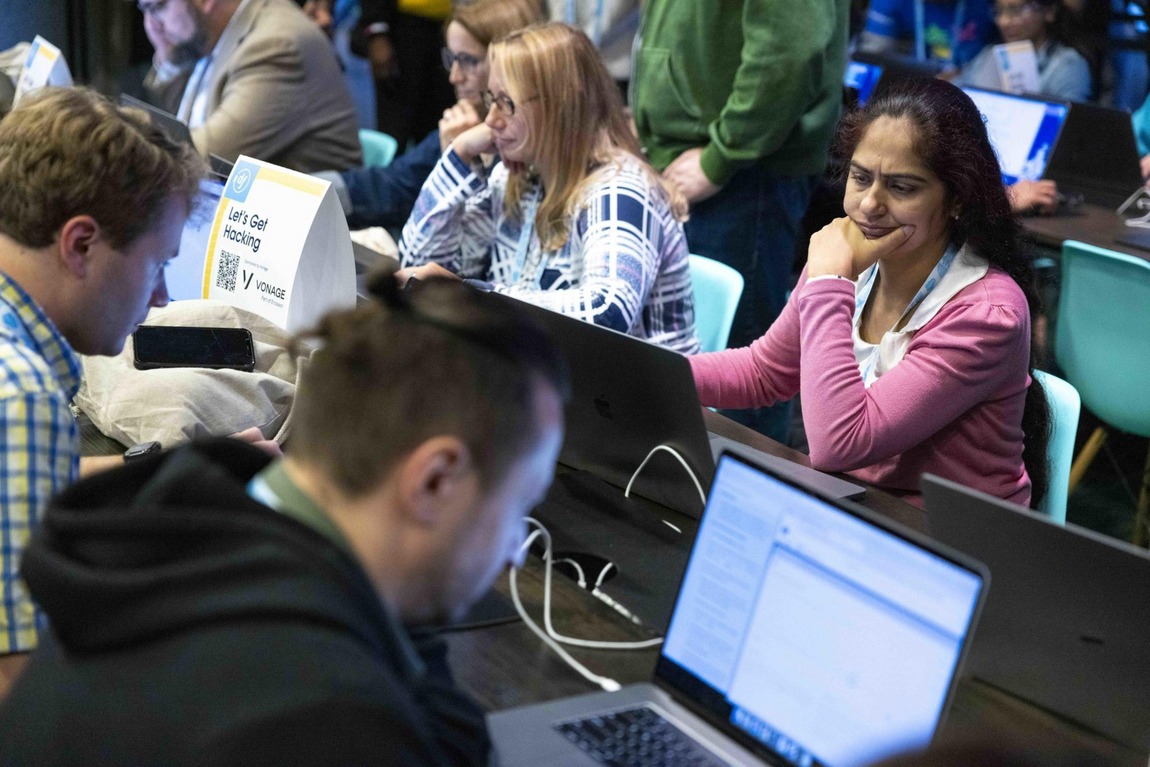 People are seated at a table, focused on laptops. A sign reads &quot;Let's Get Hacking.&quot; The setting appears to be a tech event or hackathon.