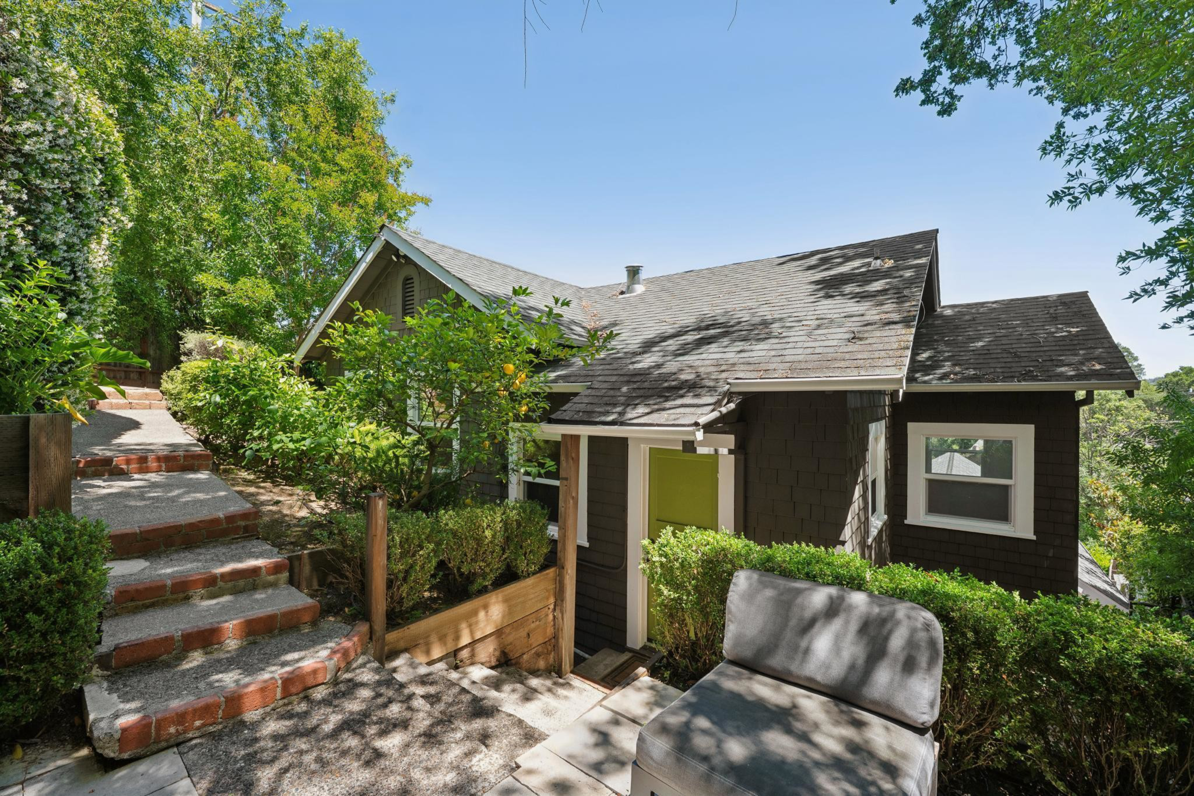 A small dark brown house with a green door sits beside a brick stairway surrounded by lush greenery and a gray cushioned outdoor chair.