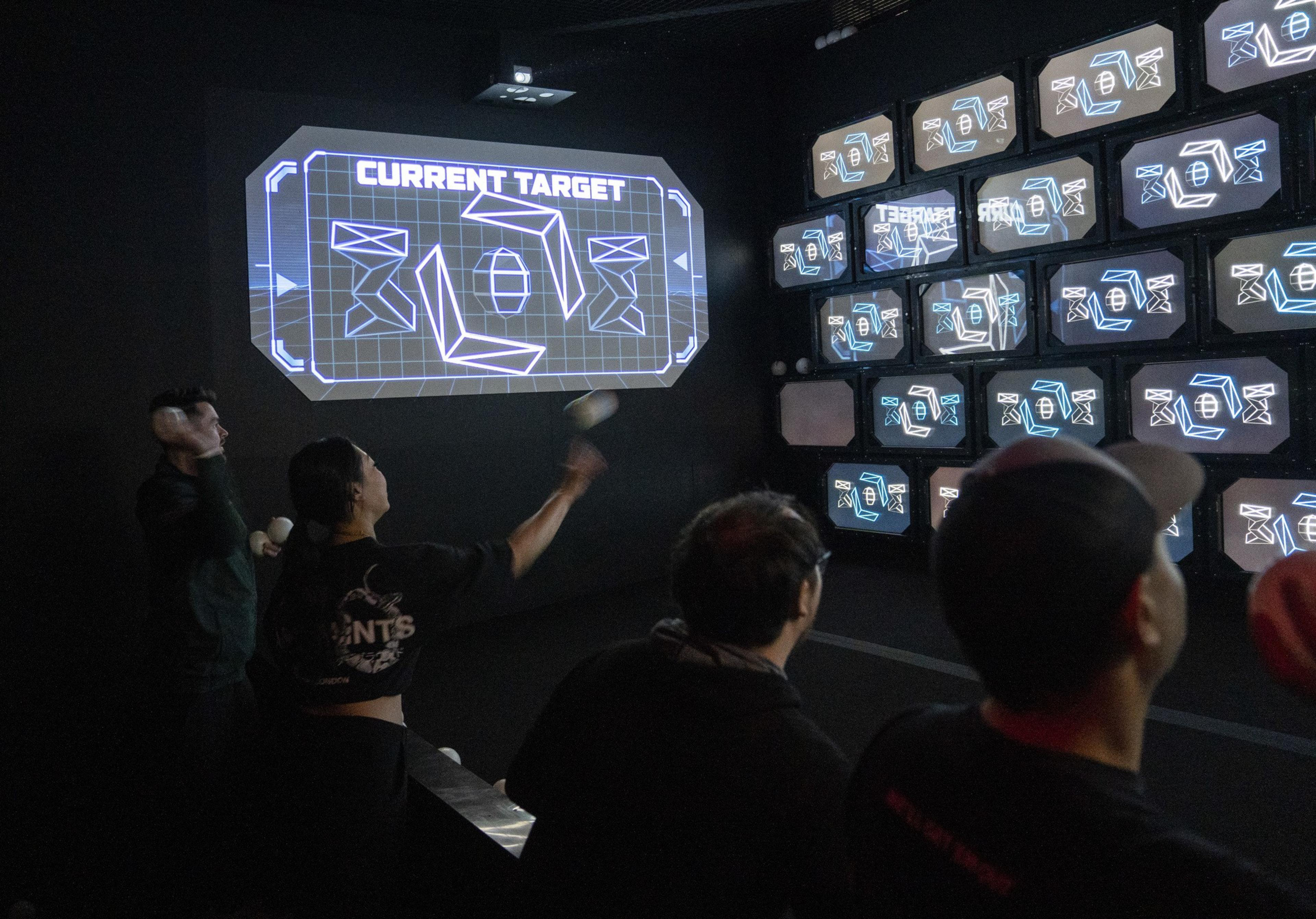 A group of people is throwing balls at a wall of screens displaying geometric shapes. The main screen reads "CURRENT TARGET," indicating a game or activity.