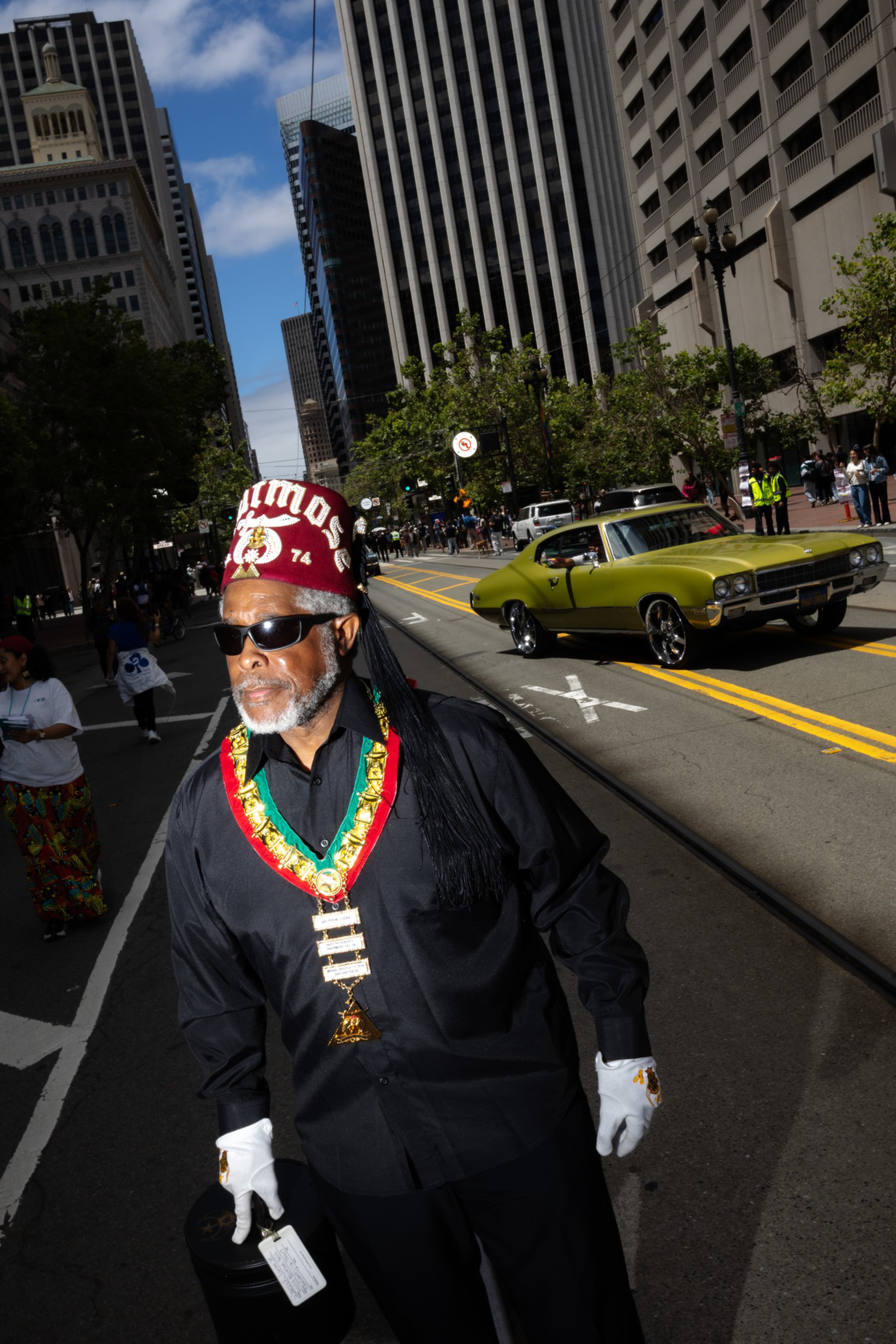 A man wearing dark sunglasses, a red fez hat, black outfit with white gloves, and a colorful medallion stands on a city street near a green vintage car.