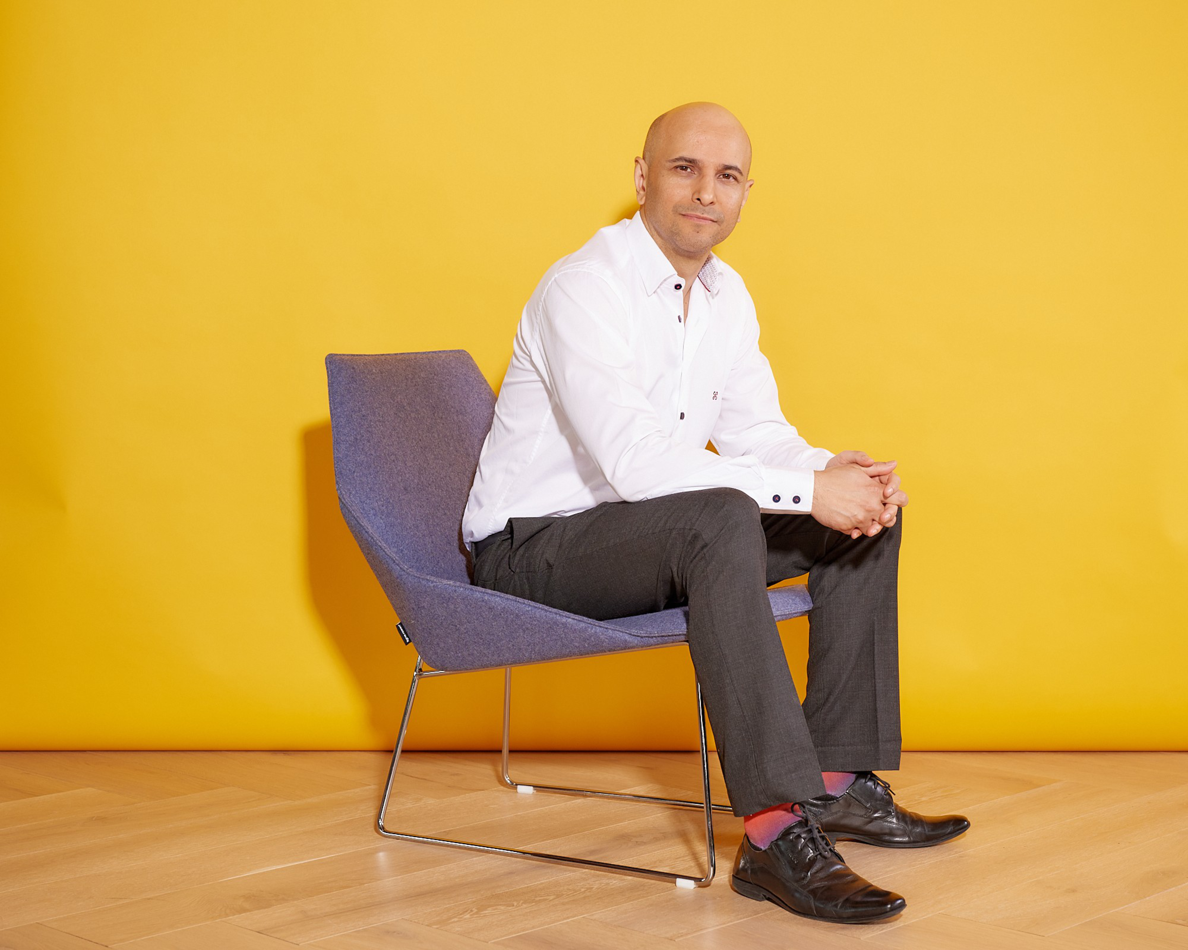 A bald man in a white shirt and dark pants sits on a modern blue chair against a vibrant yellow background, hands clasped and looking forward.