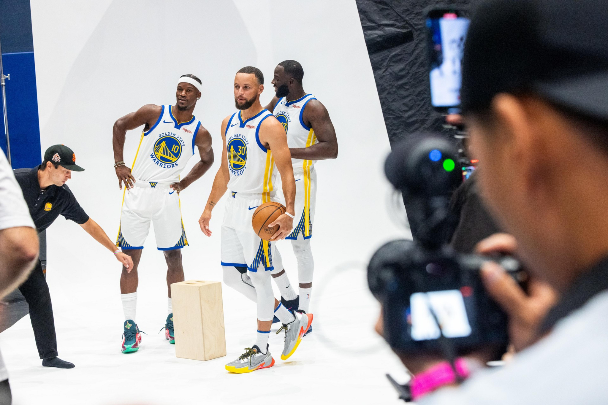 Three Golden State Warriors players in white uniforms pose for a photoshoot while a photographer captures their image.