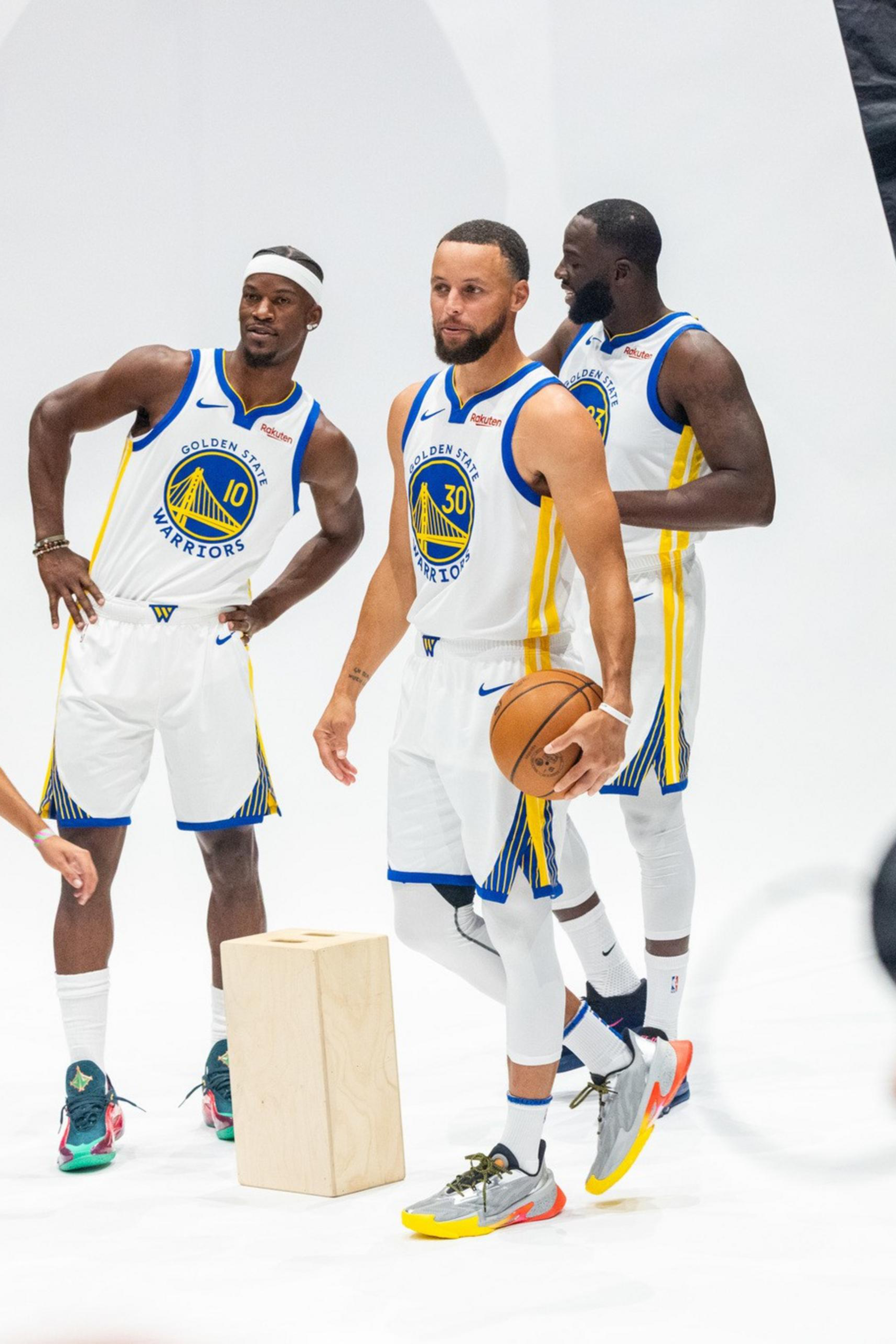 Three Golden State Warriors players in white uniforms pose for a photoshoot while a photographer captures their image.