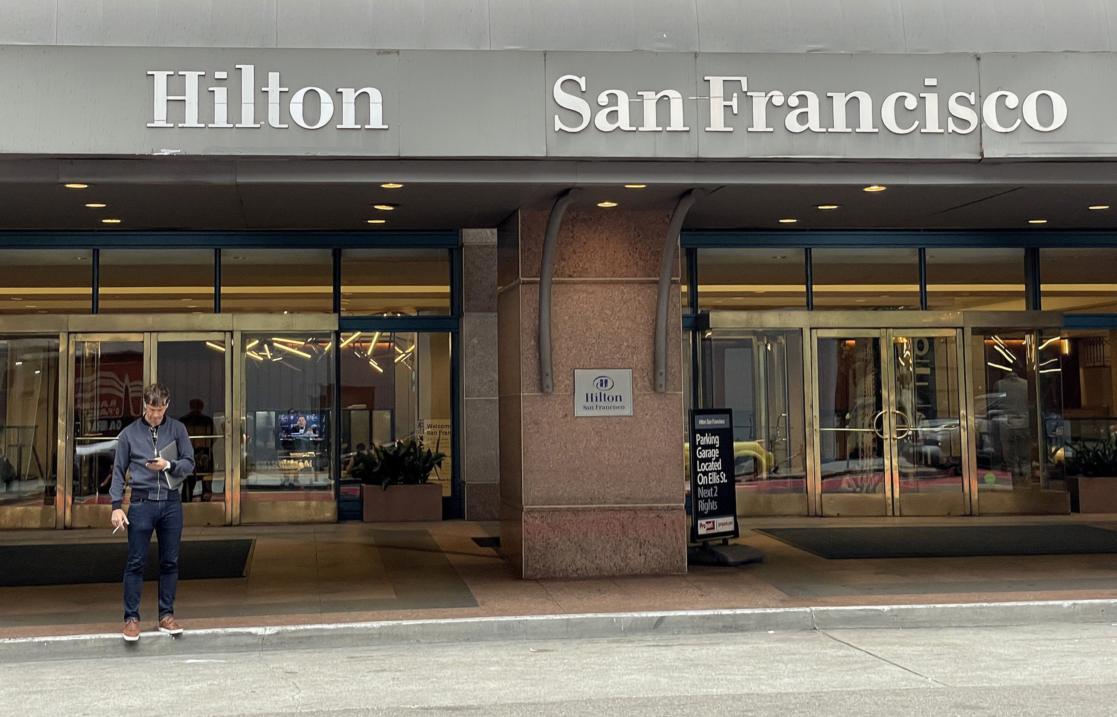 The image shows the entrance of the Hilton San Francisco with a man looking at his phone while standing on the sidewalk in front of the glass doors.