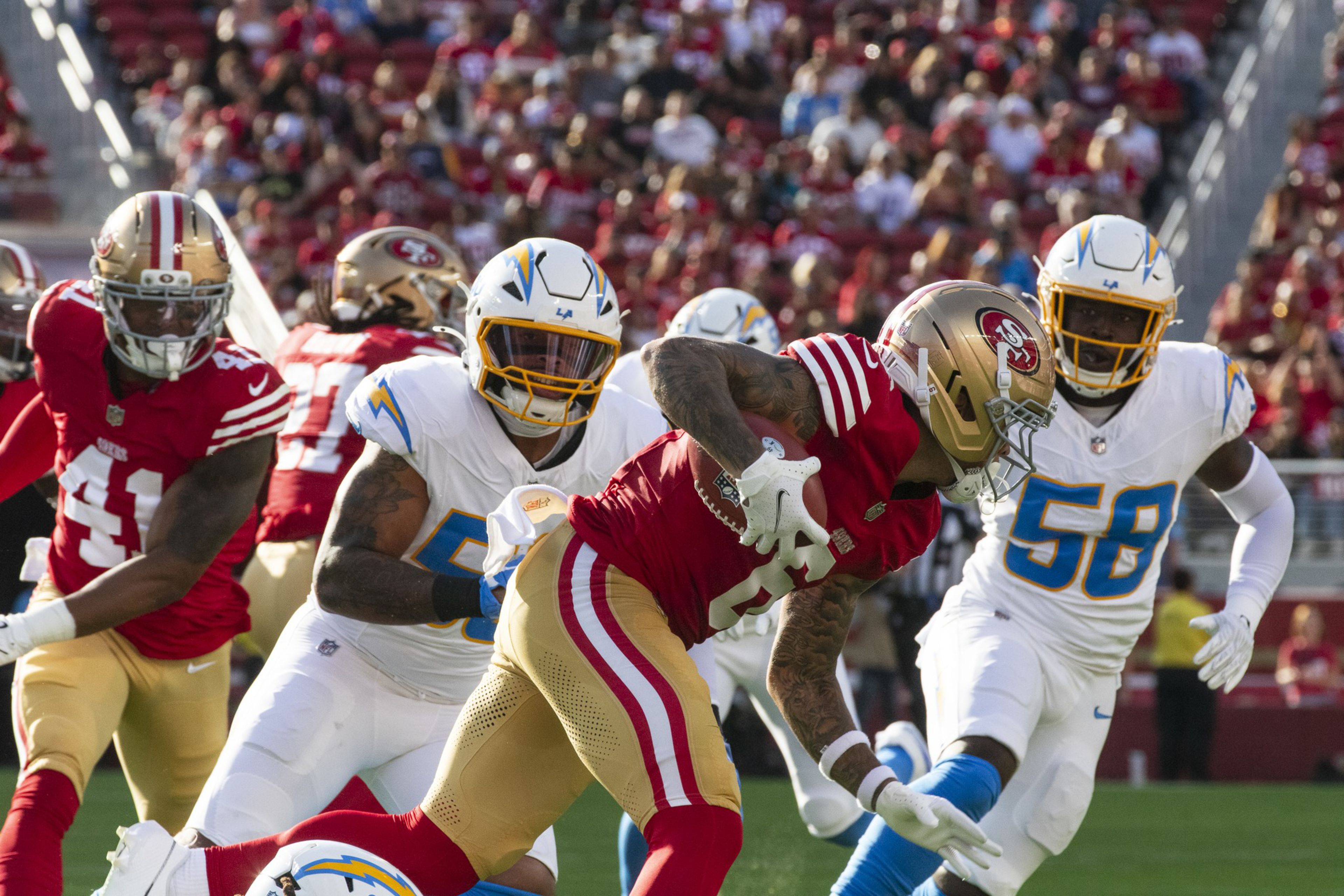 A San Francisco 49ers player runs with the ball while Los Angeles Chargers defenders rush to tackle him during a football game.