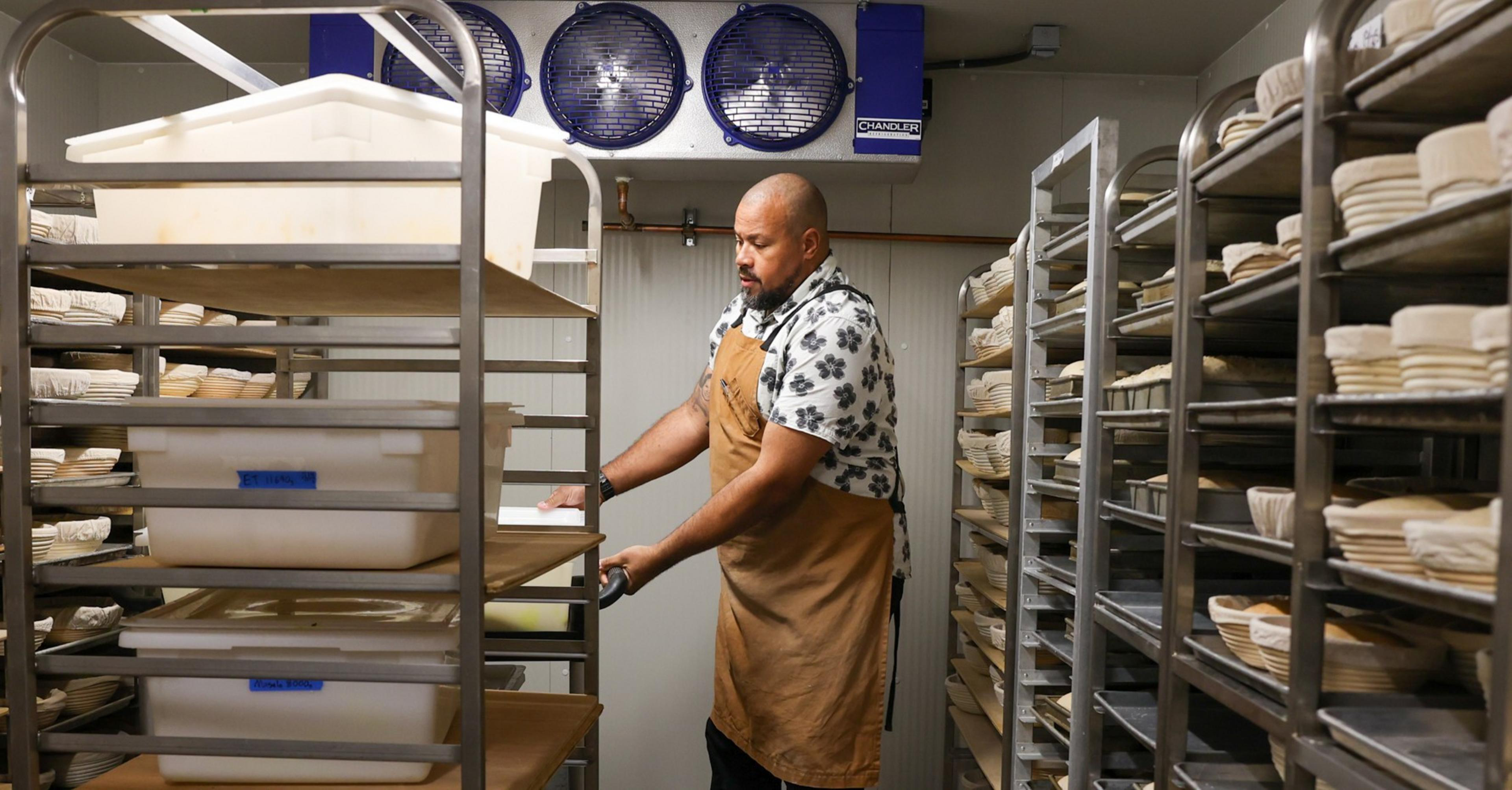 A shaven-headen man stands between tall metal shelf racks laden with dough and bread.