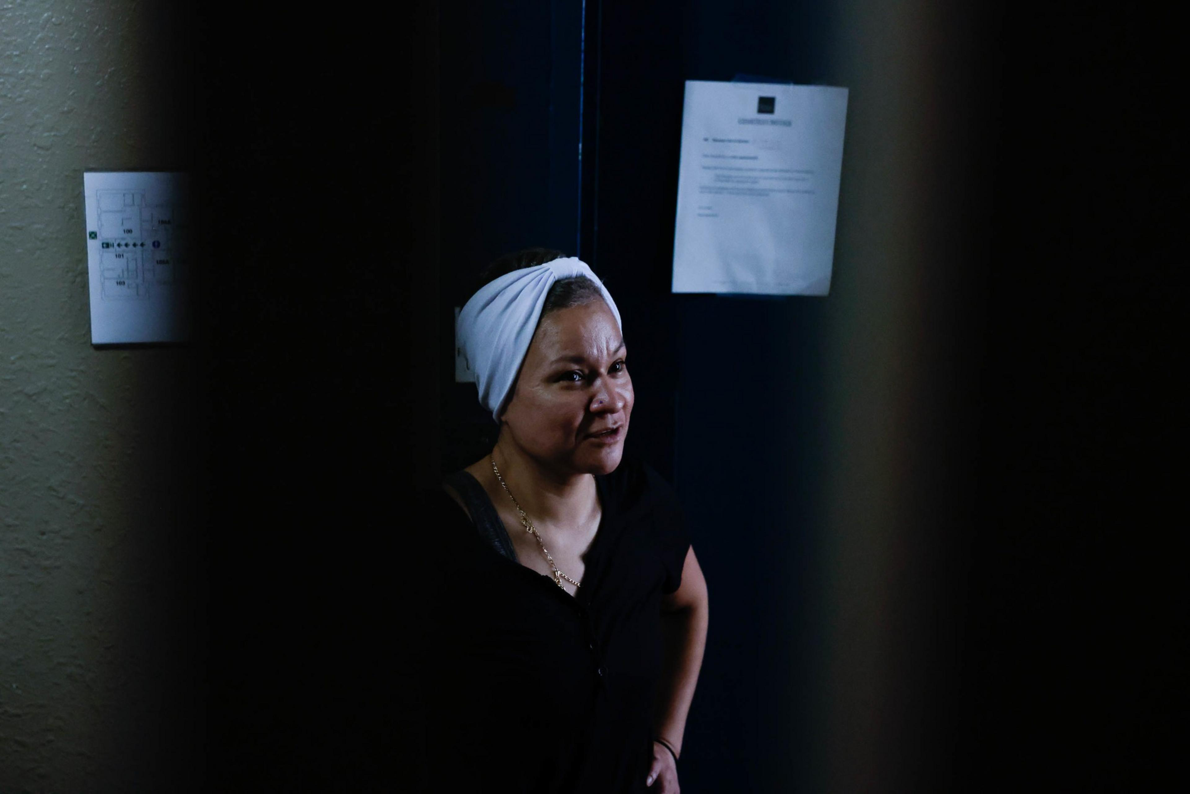 A woman with a white head wrap and black top looks pensively off to the side in a dimly lit corridor with papers on the wall.