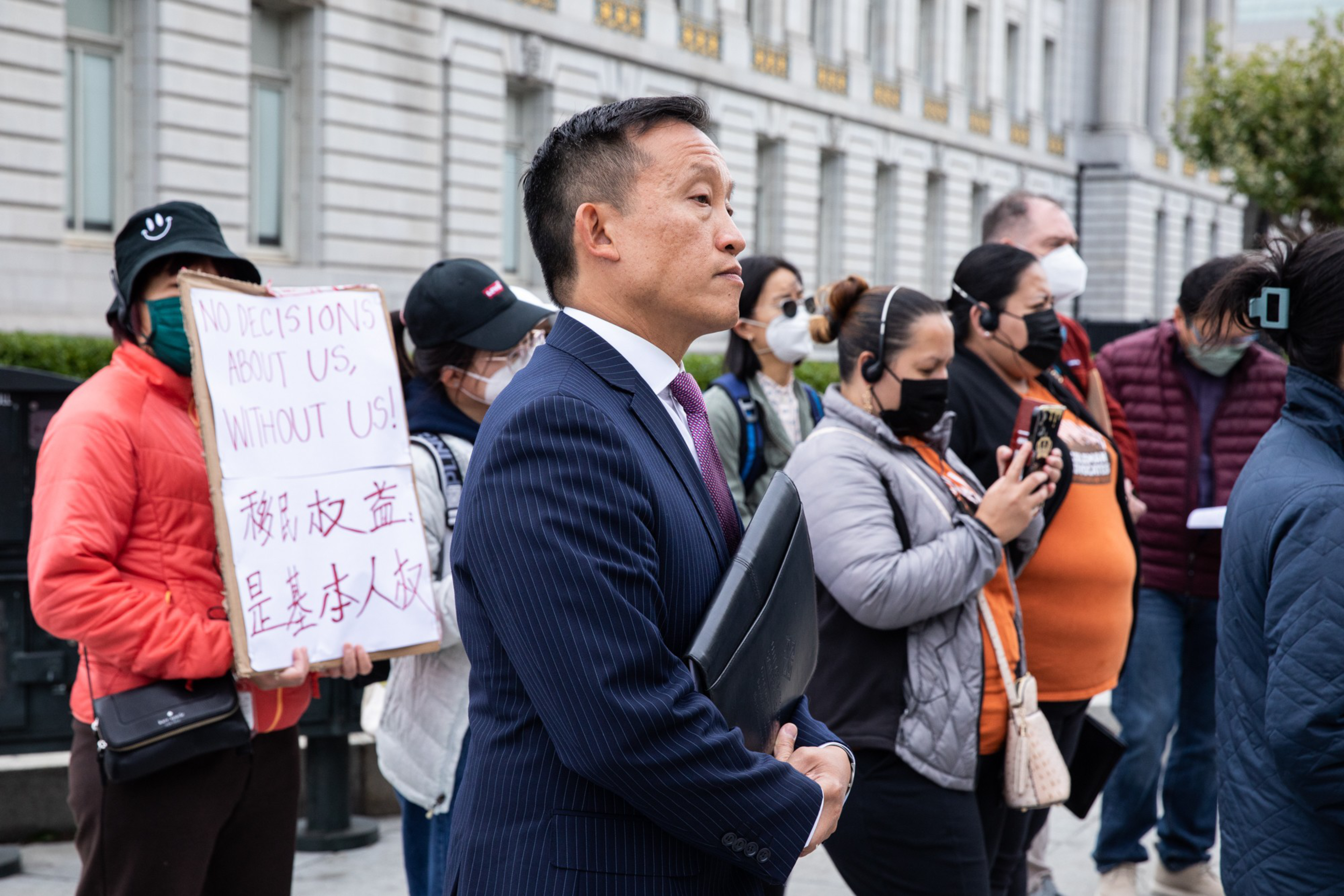 A man in a suit holds a folder, standing among a group of people, some with masks. One person holds a sign saying, "No decisions about us, without us!"
