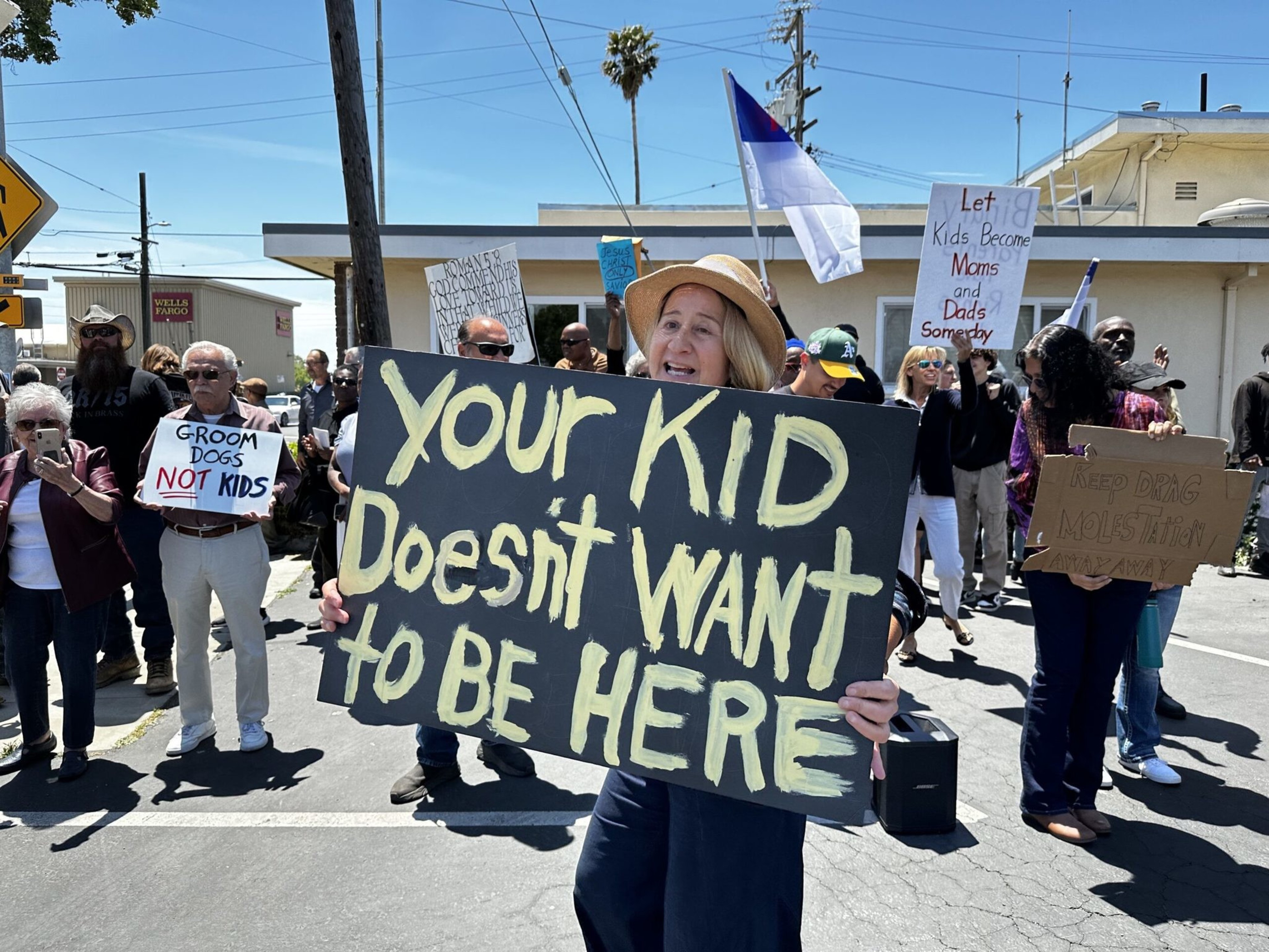 A group of protesters hold signs, including one reading "YOUR KID Doesn't WANT to BE HERE" and others with messages about kids, grooming, and drag.