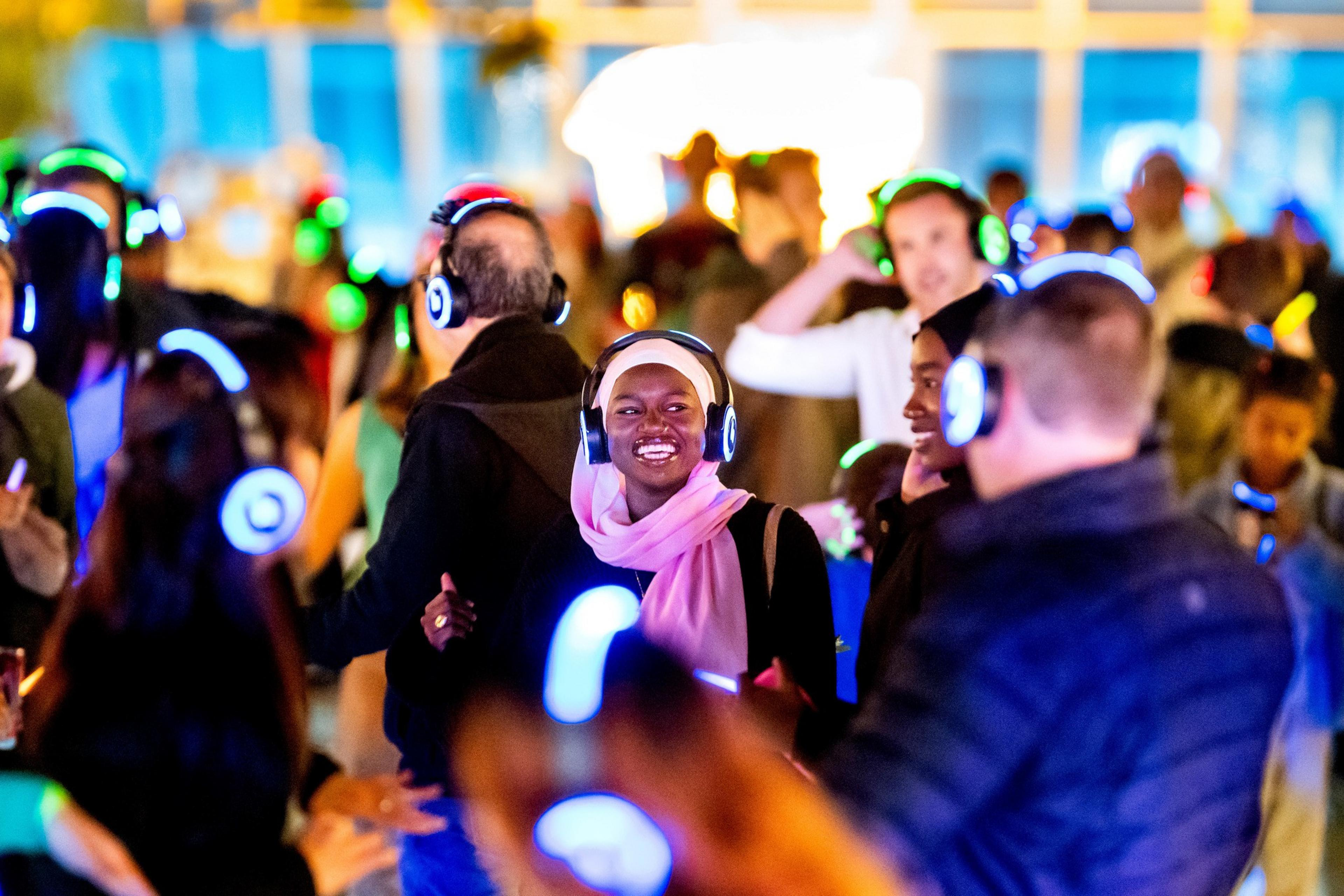 A group of people is enjoying a silent disco, wearing glowing, colorful headphones and smiling. The background is blurred with vibrant lights.