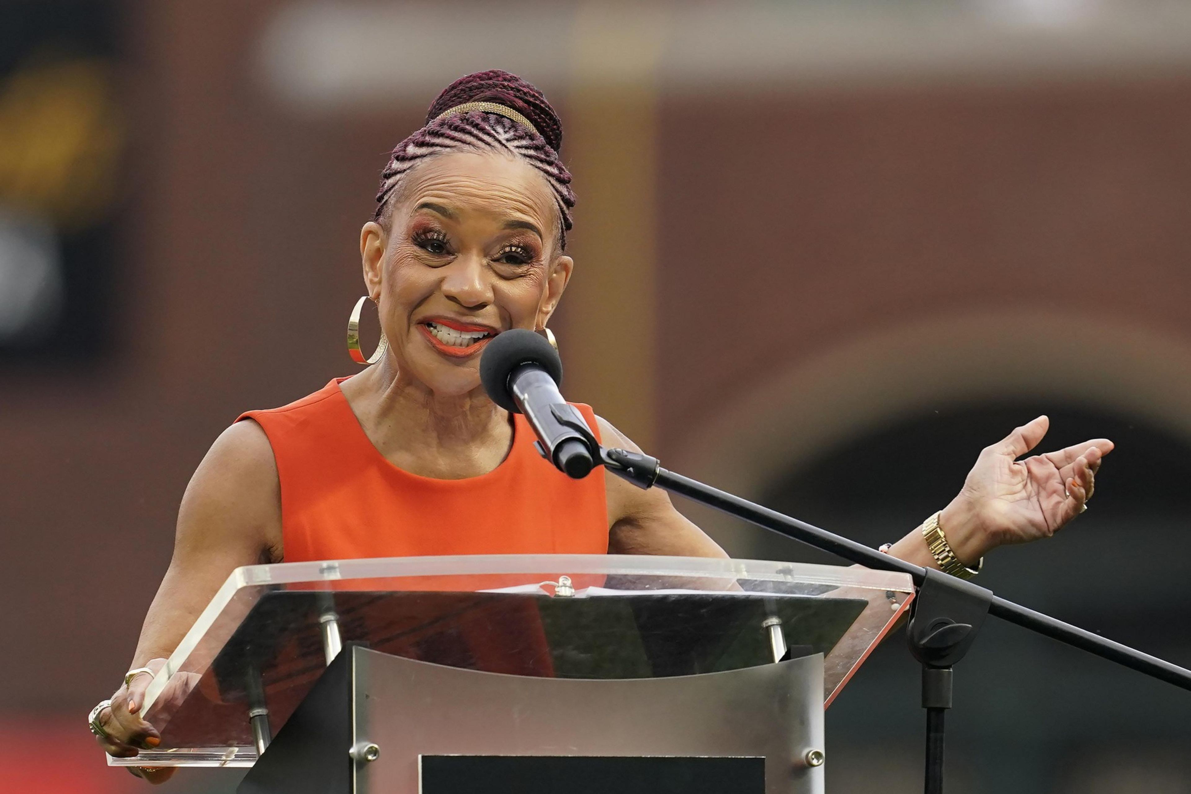 A person with braided hair wearing an orange outfit speaks animatedly at a podium. They gesture with one hand while smiling and speaking into a microphone.