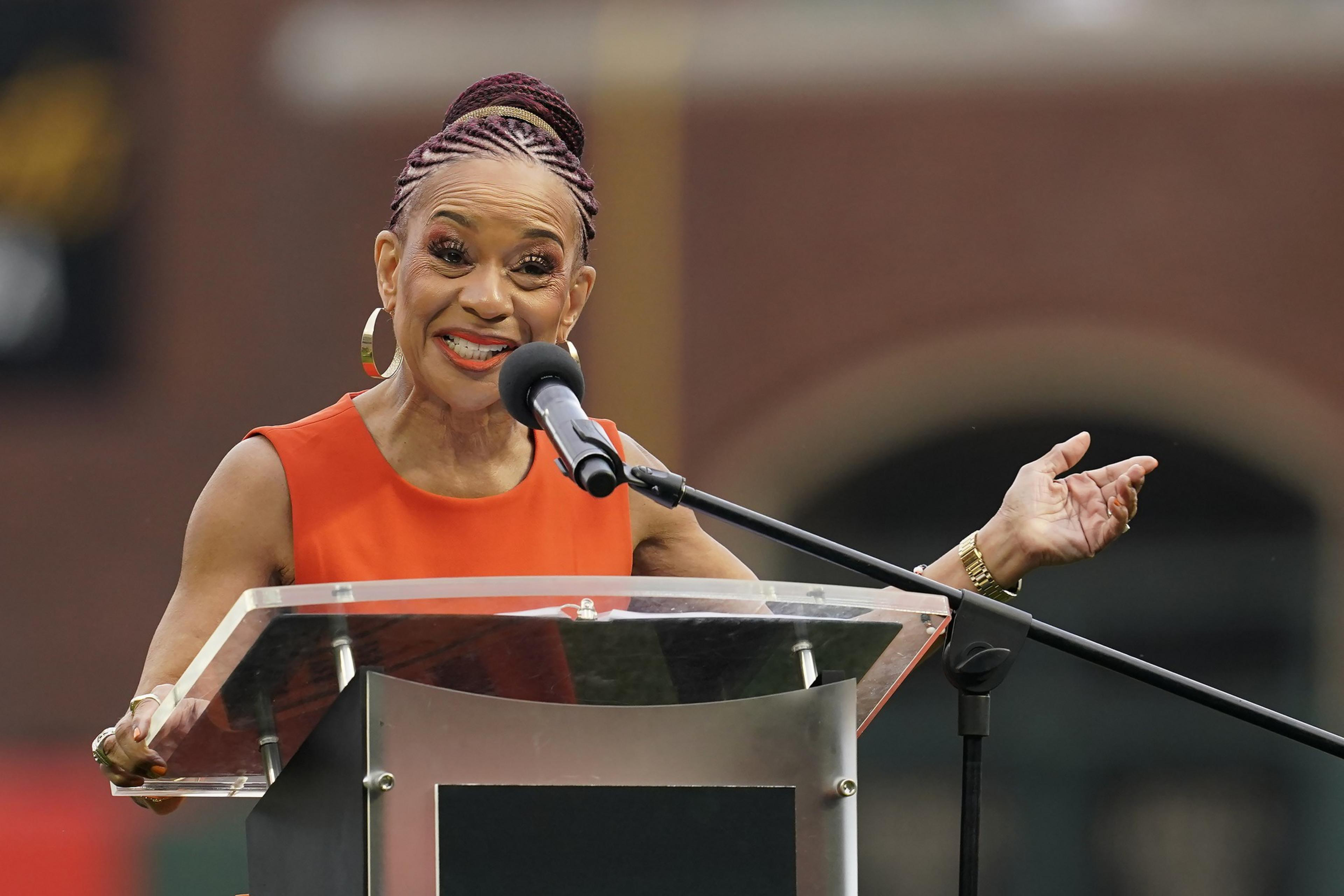 A person with braided hair wearing an orange outfit speaks animatedly at a podium. They gesture with one hand while smiling and speaking into a microphone.