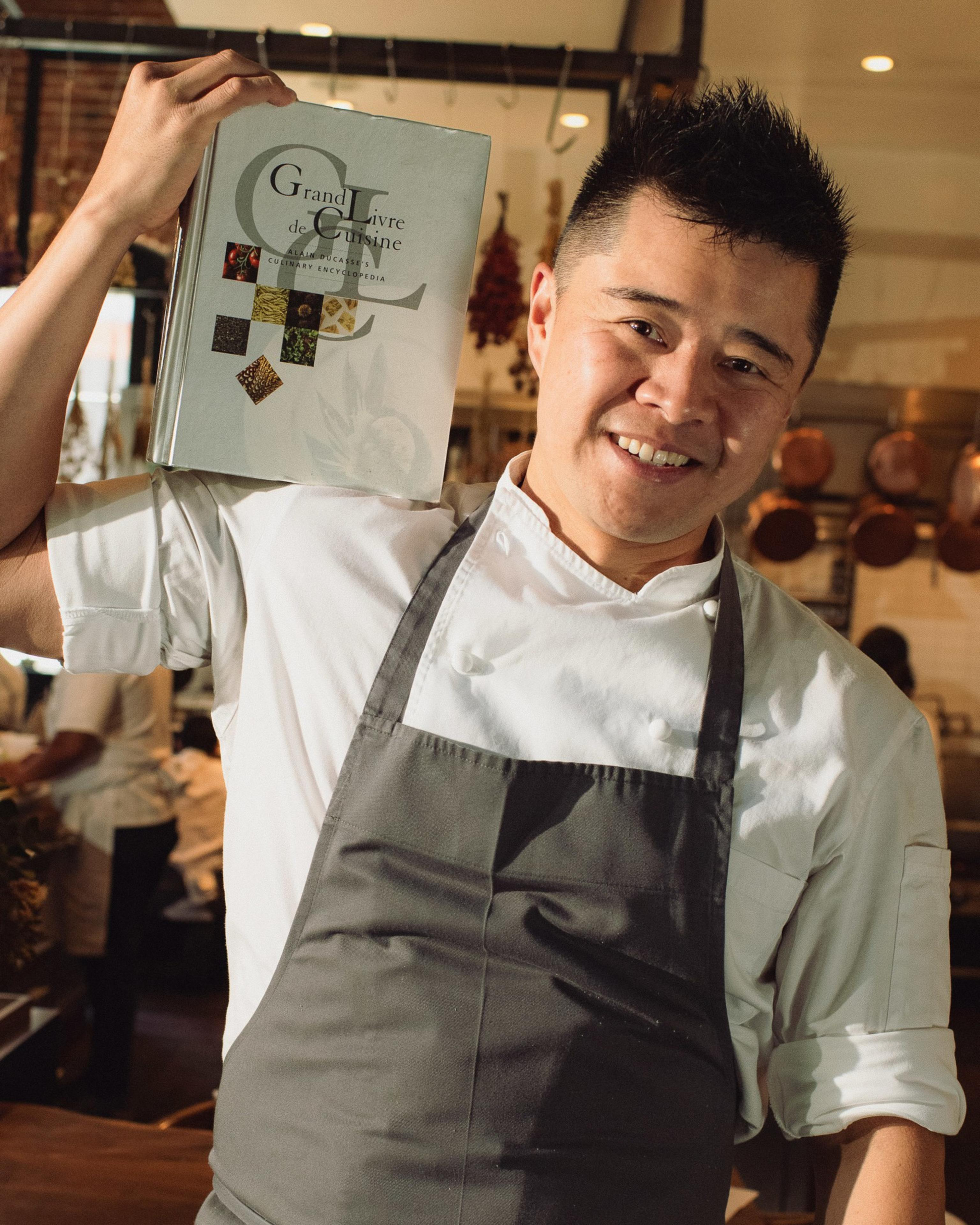 A smiling chef in a white coat and gray apron holds a large cookbook titled "Grand Livre de Cuisine" on his shoulder in a warm kitchen setting.