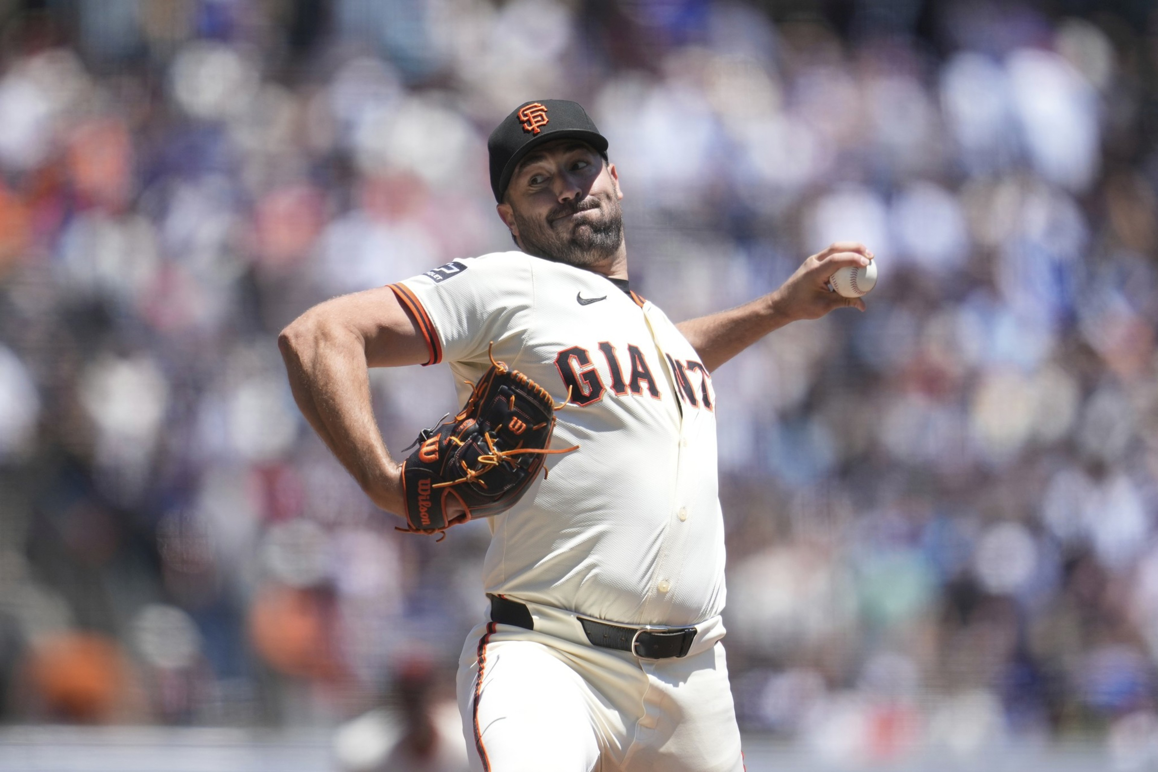 A baseball player in a white Giants uniform and black cap is pitching in a stadium. He's in motion, with his arm extended holding a baseball.