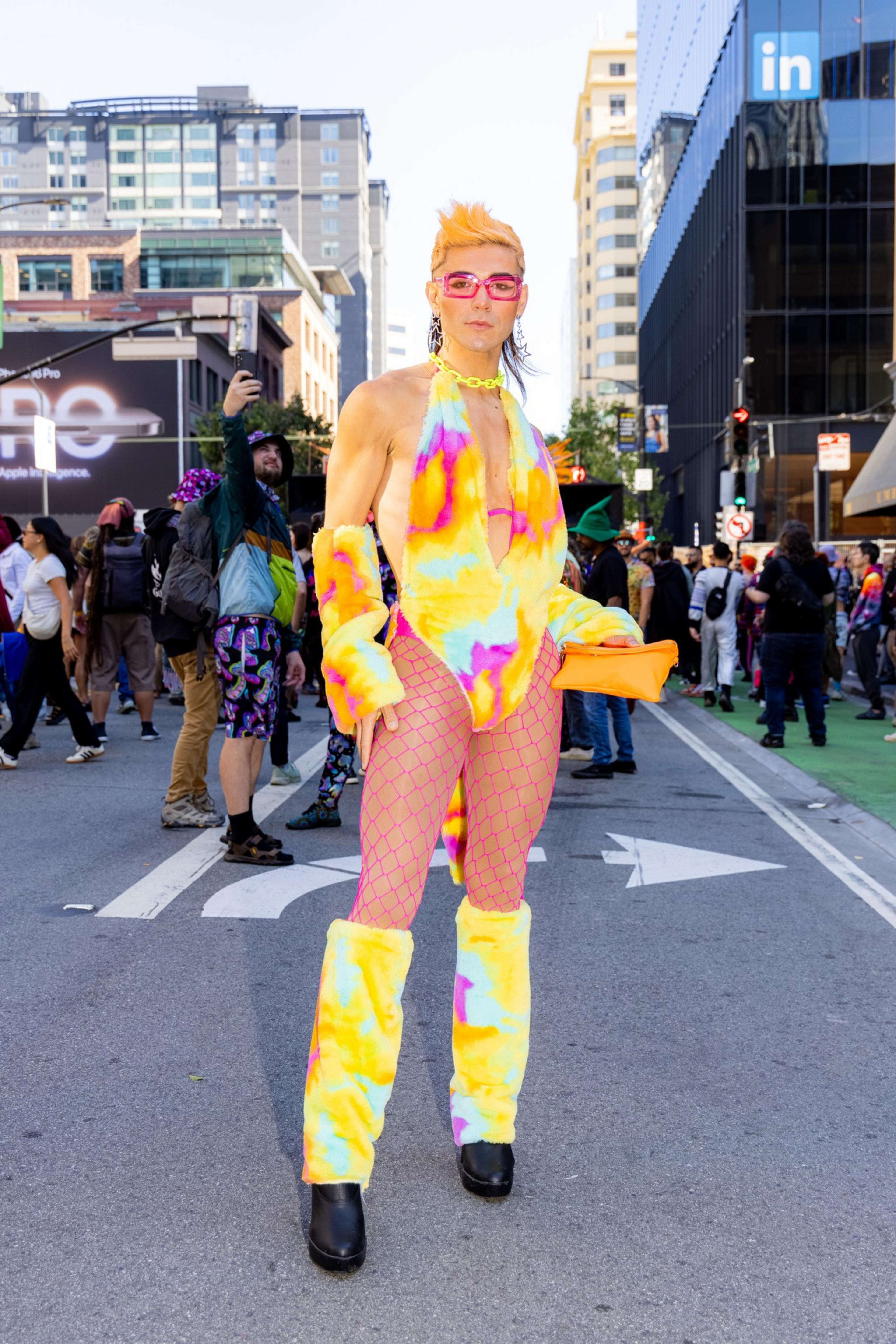A person stands confidently in the street, dressed in a colorful, tie-dye bodysuit with fishnet stockings, furry accessories, and pink glasses, amidst a city crowd.