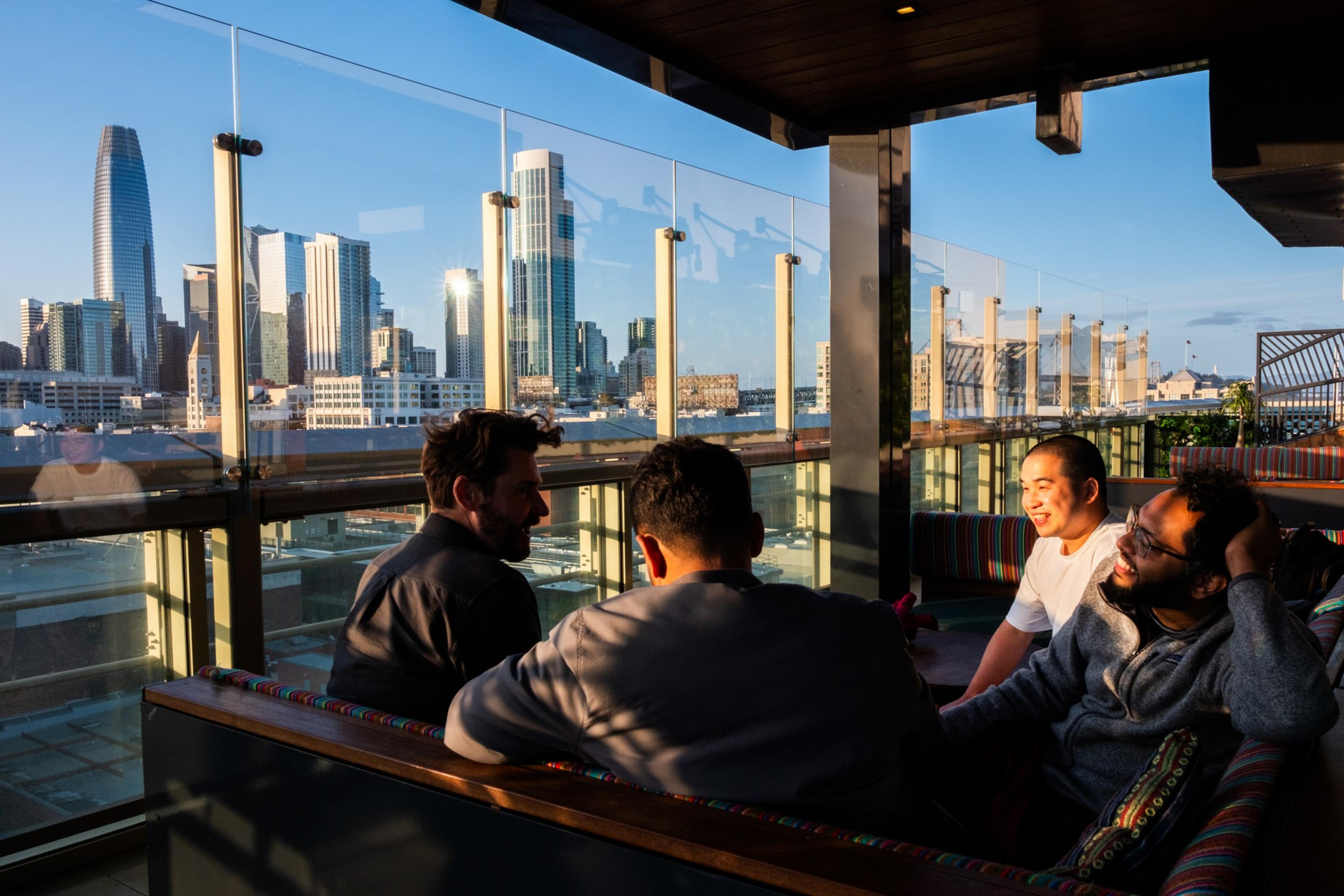 Four people are sitting on a rooftop terrace with city skyscrapers in the background, chatting and smiling in the warm, golden light of sunset.