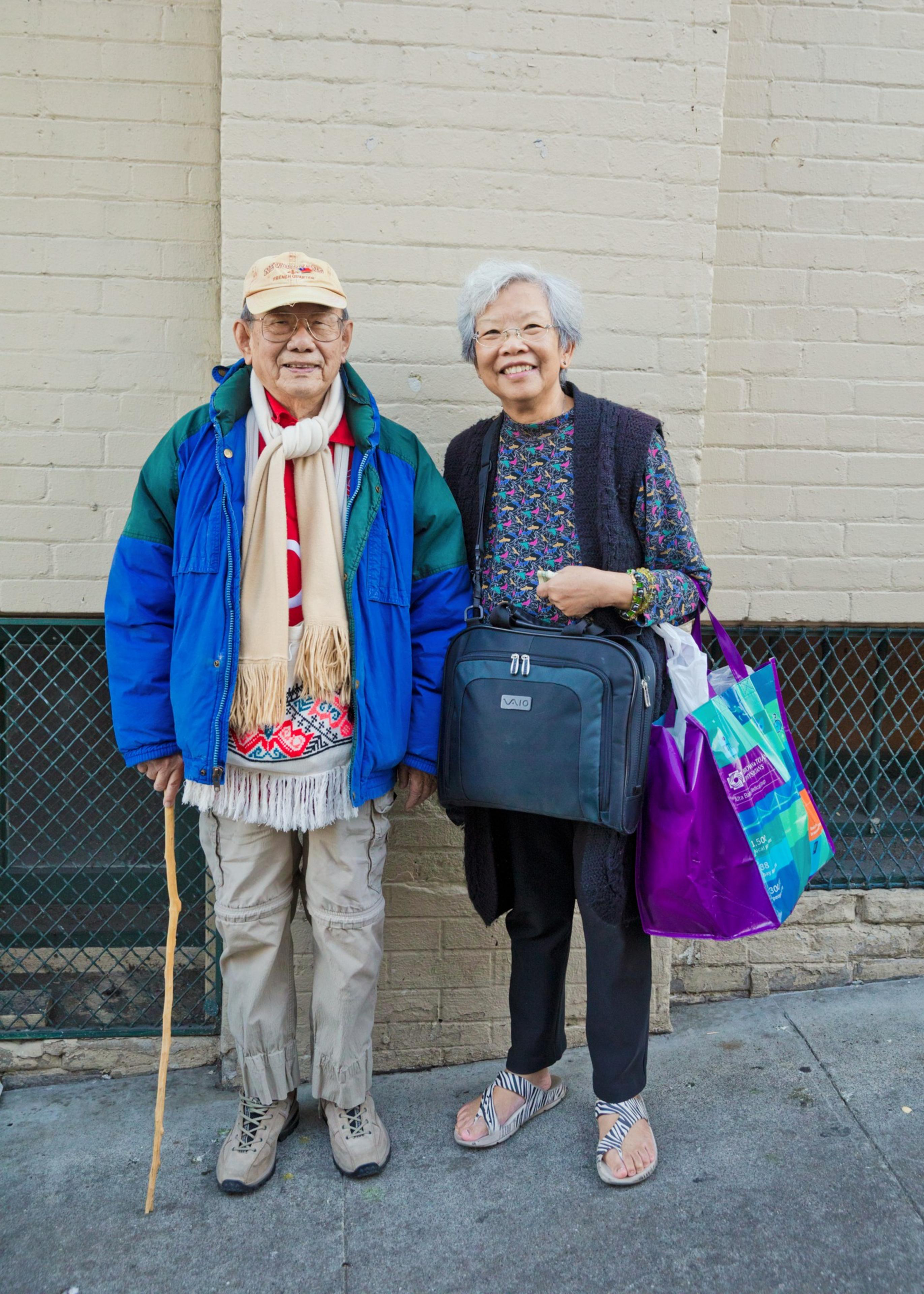 An elderly man and woman stand side by side, smiling, with the man holding a wooden cane and the woman carrying multiple bags.