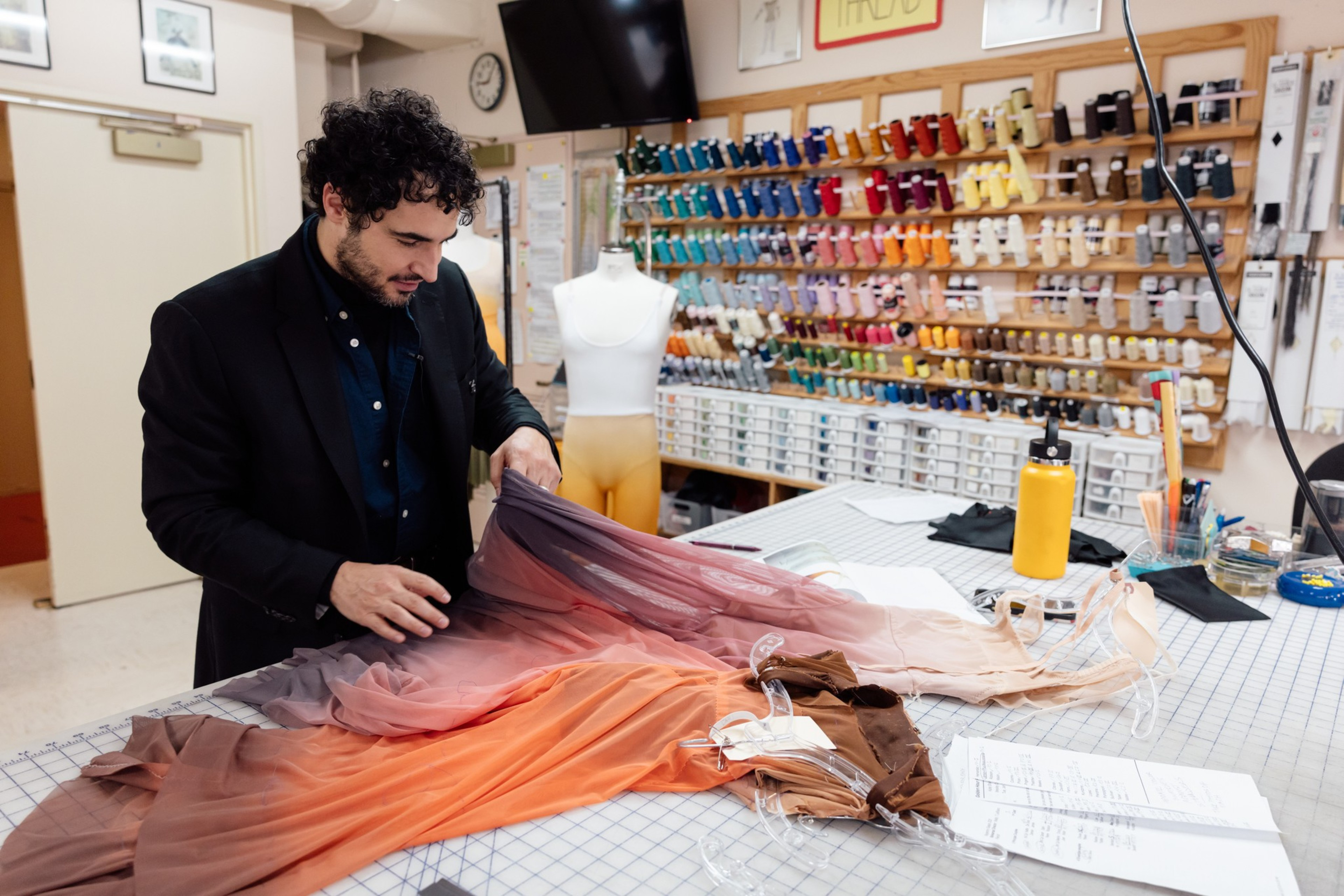 A man examines colorful fabric on a table in a sewing room. The wall holds numerous spools of thread in various colors, and a dress form stands nearby.